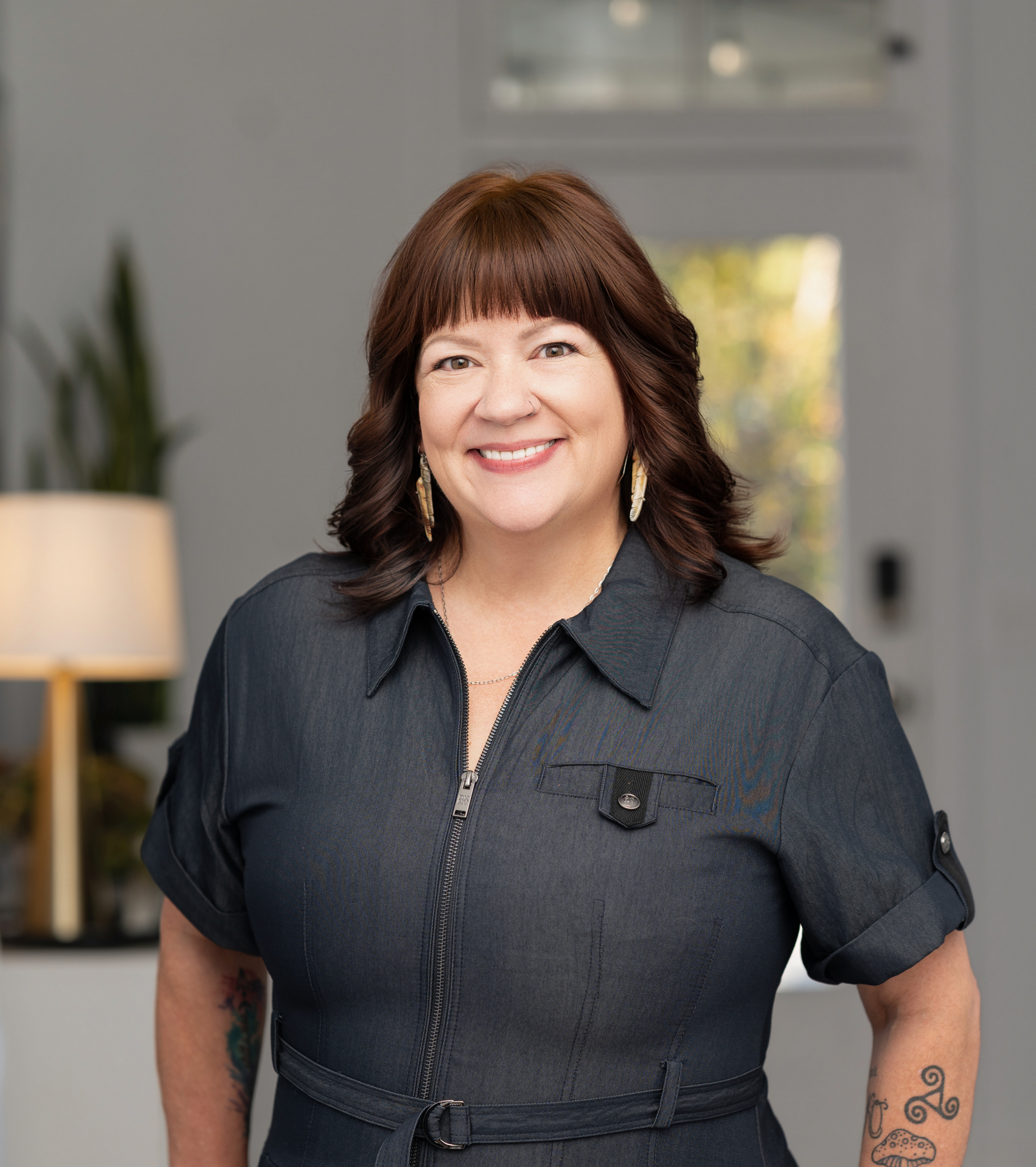 A smiling white woman with auburn hair and bangs, wearing a medium blue denim dress.