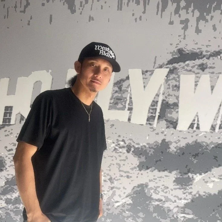 A man wearing a black cap and black T-shirt standing in front of a mural with the Hollywood sign in the background.