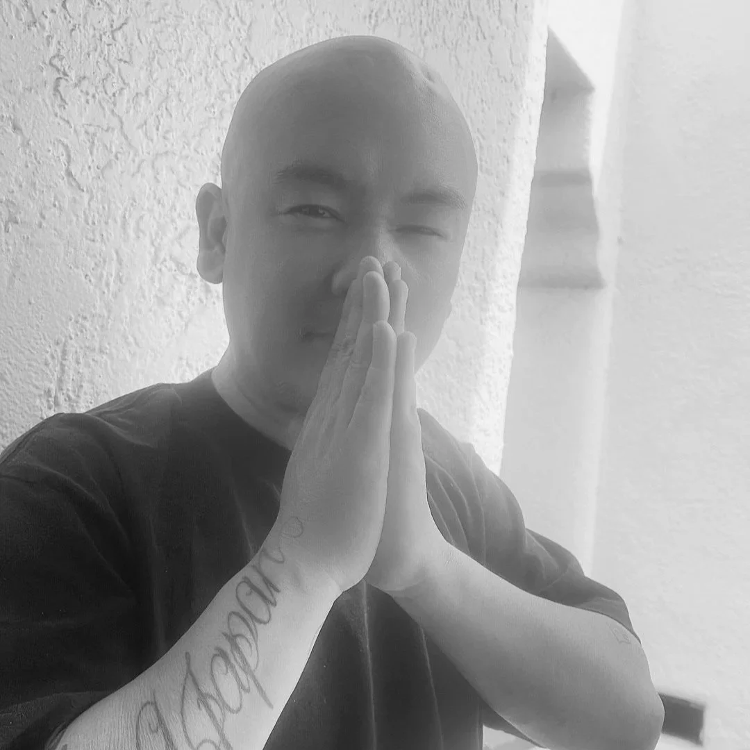 Black and white photo of a bald man with tattoos on his arms, holding his hands together near his face in a prayer-like gesture, standing against a textured wall.
