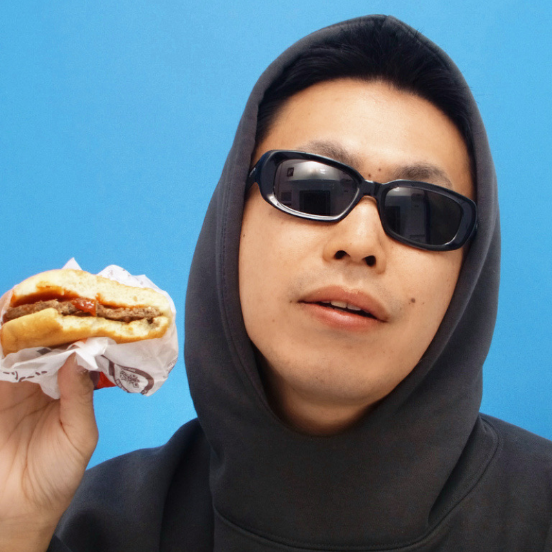 A young man wearing sunglasses and a gray hooded sweatshirt holding a cheeseburger with lettuce, tomato, and meat against a solid blue background.