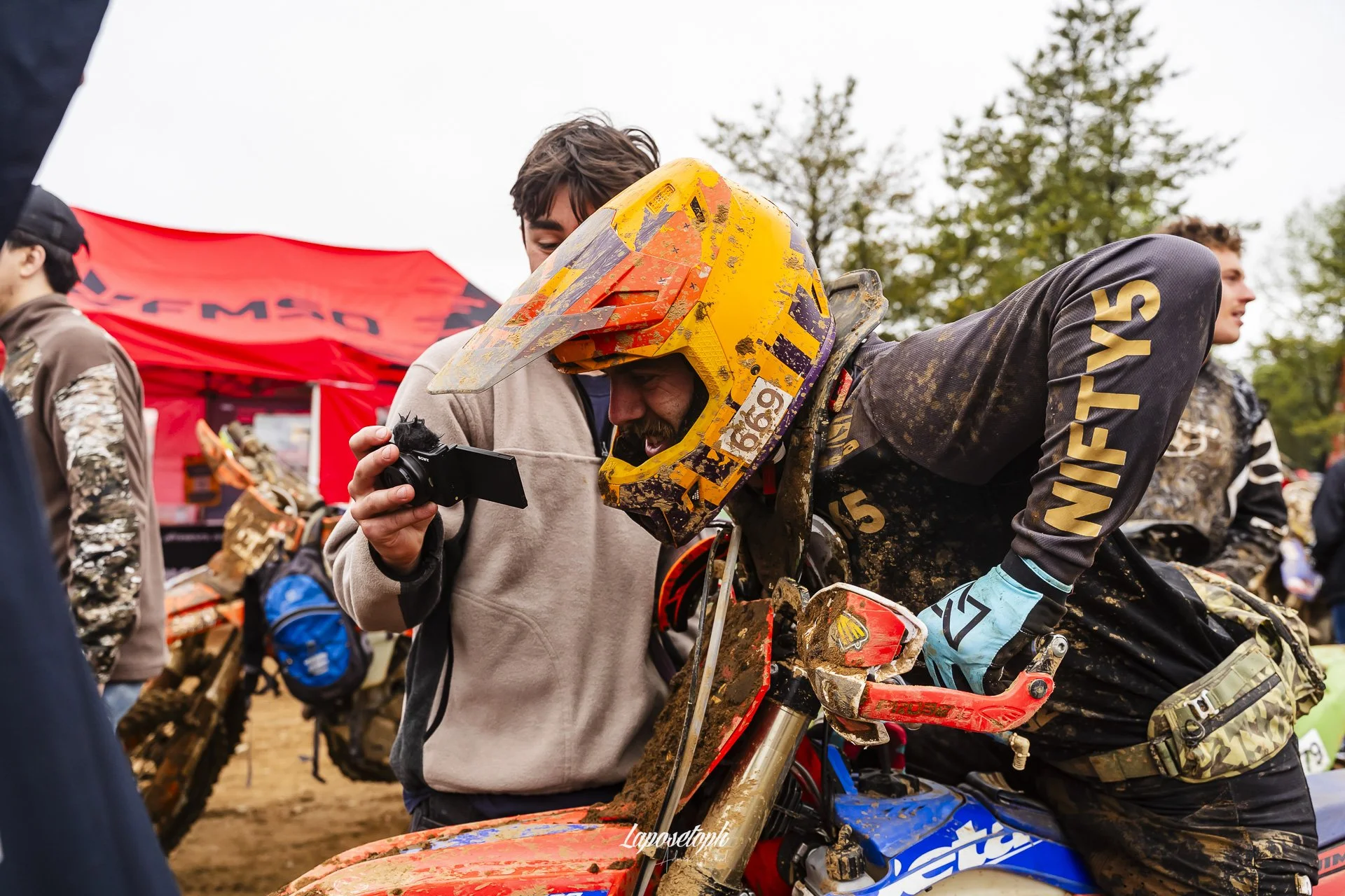 A motocross rider in muddy gear and a bright yellow helmet leaning on his dirt bike, with other racers and a red tent in the background.