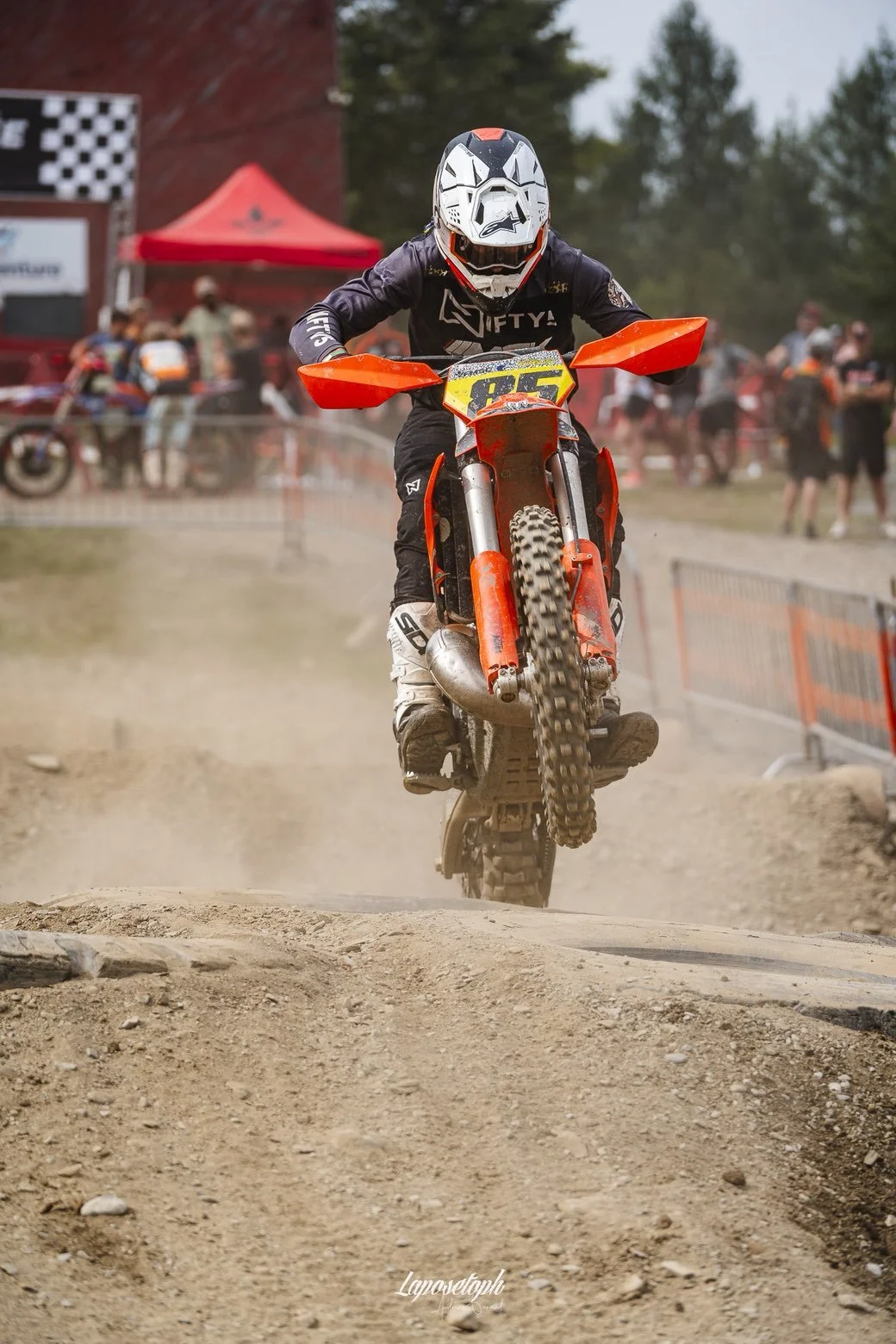 Motocross rider wearing helmet and gear, riding a dirt bike through a dirt track with dust and spectators in the background.