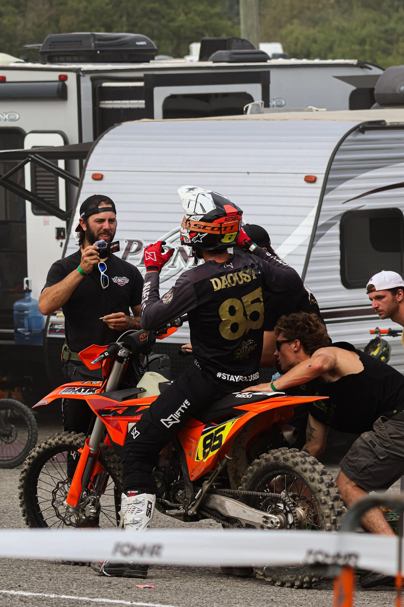 A motocross rider wearing a helmet and black gear with the number 85, sitting on an orange KTM dirt bike. Several people surround him, some appear to be talking or assisting, and one is holding a microphone. In the background, there are trailers and 