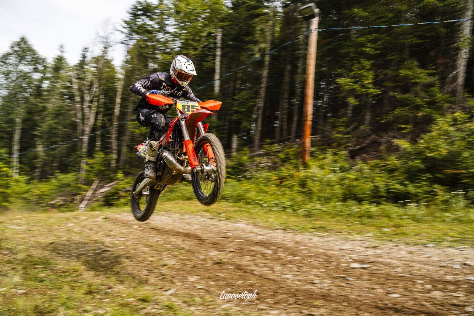 Motocross rider jumping over a dirt track in an outdoor forest setting, wearing a helmet and black gear, with trees in the background.