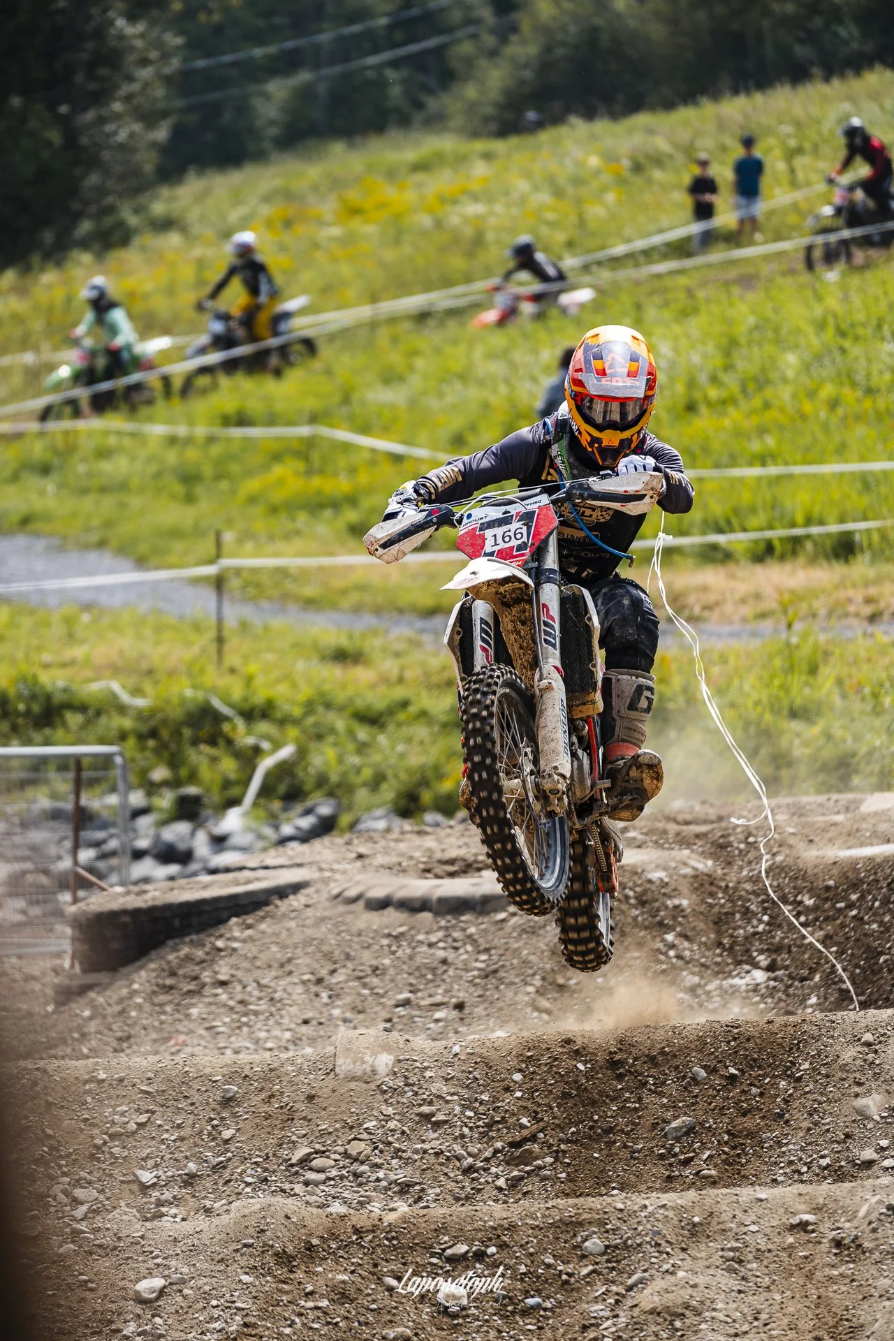 A motocross rider wearing a helmet and protective gear jumping over a dirt mound on a motocross track, with dirt flying from the tires, in a rural outdoor setting.