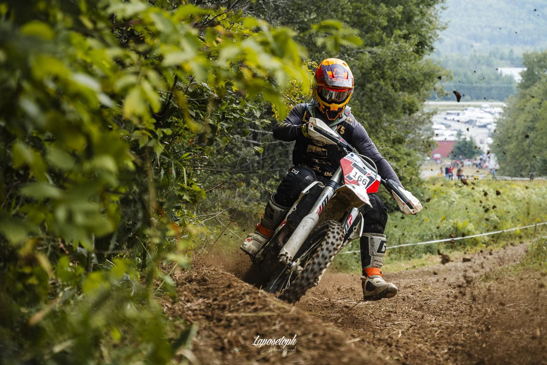 A motocross rider wearing a full-face helmet and riding gear, navigating a dirt trail surrounded by greenery, with a background of trees, vehicles, and spectators.