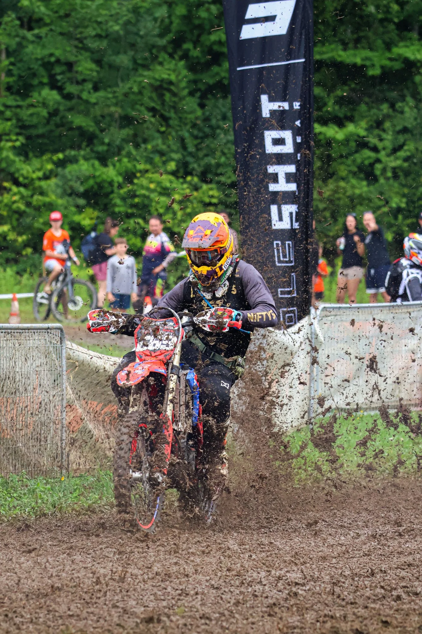 A motocross rider wearing a yellow helmet and black gear riding through a muddy track, with spectators and a vertical banner in the background.