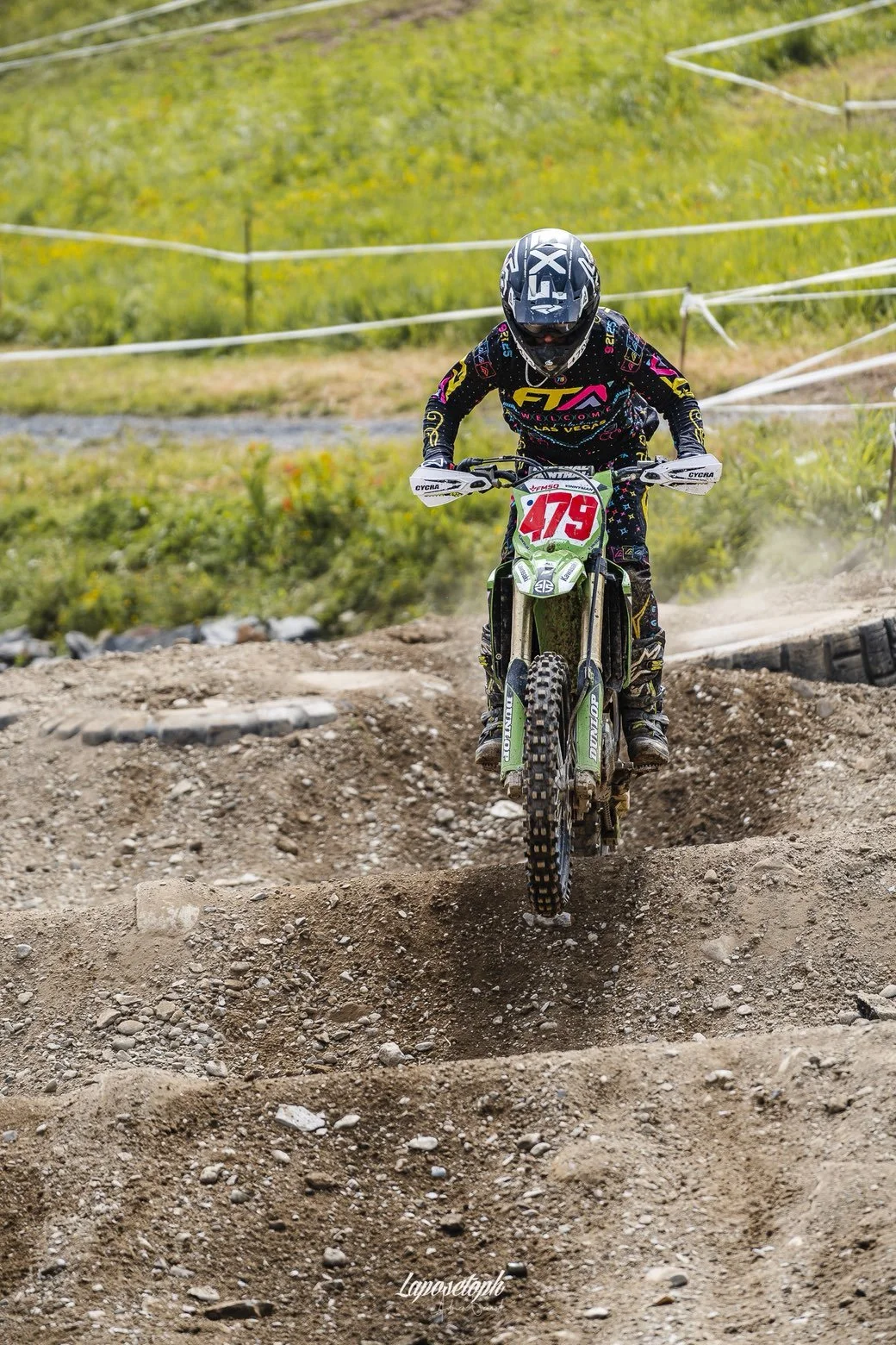 A motocross rider wearing a helmet and colorful gear riding a dirt bike over a dirt ramp on an outdoor track with green grass and yellow flowers in the background.