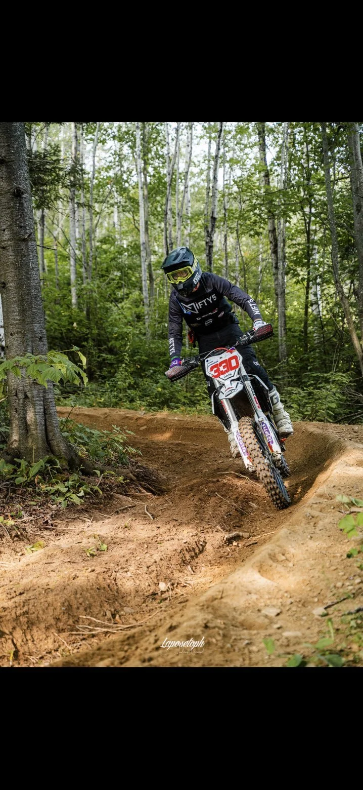 A mountain biker in a black helmet and gear rides downhill on a dirt trail in a dense forest with green trees.