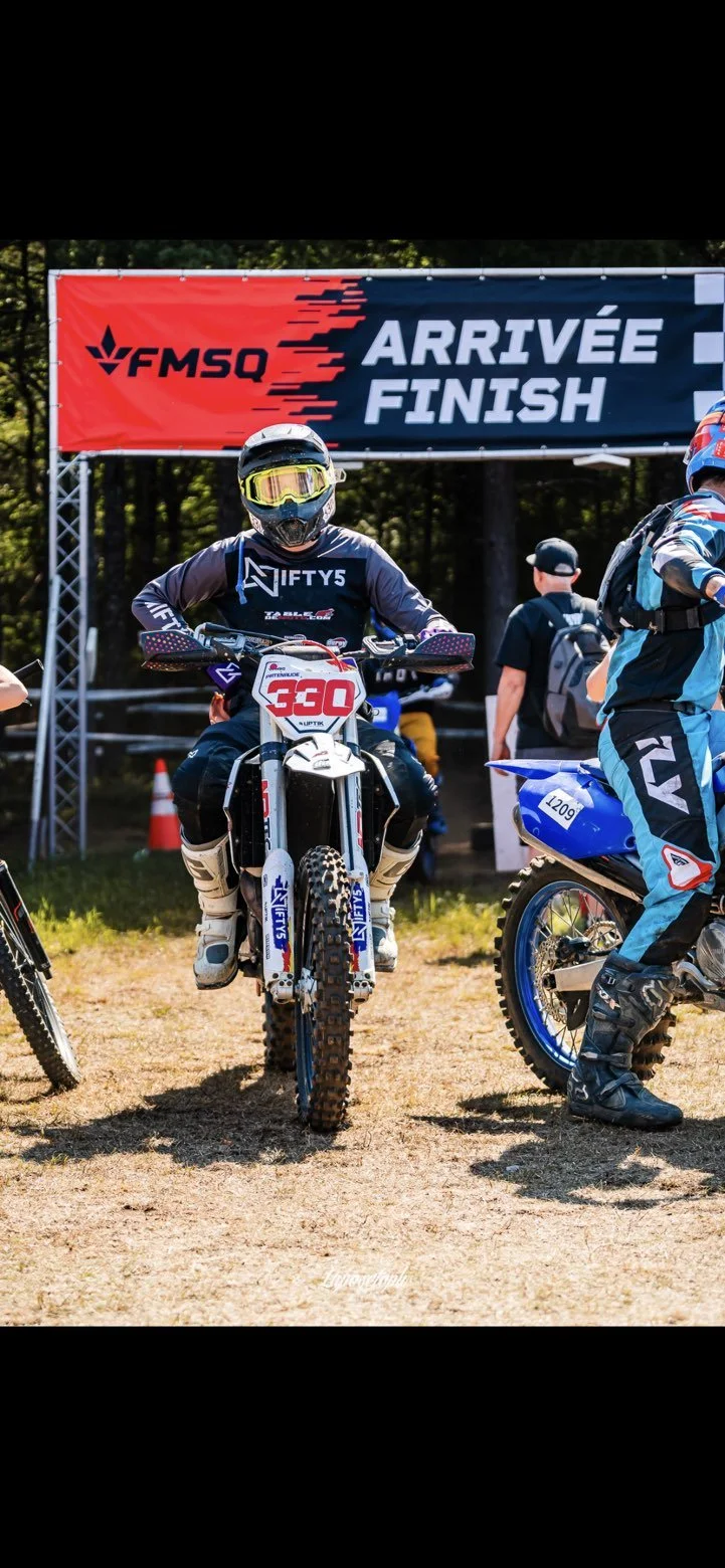 Motorcycle racer in gear at the finish line of a race, standing on a dirt track, with a banner overhead reading 'ARRIVÉE FINISH' and 'FMSQ'.