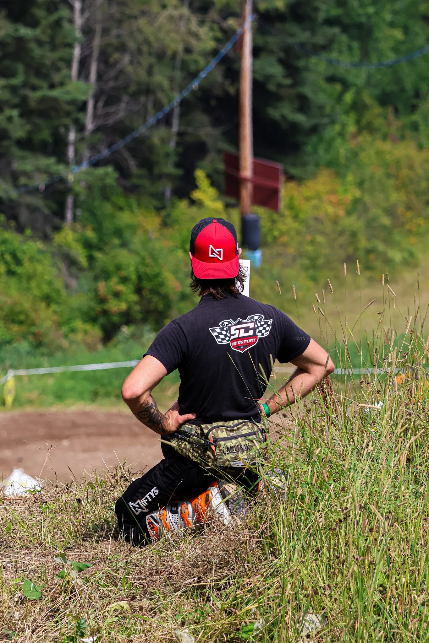 A person with dark hair, wearing a red and black cap, black shirt, camouflage fanny pack, black pants, and orange shoes, sitting on the grass with their back to the camera, outdoors on a green field with trees and utility pole in the background.