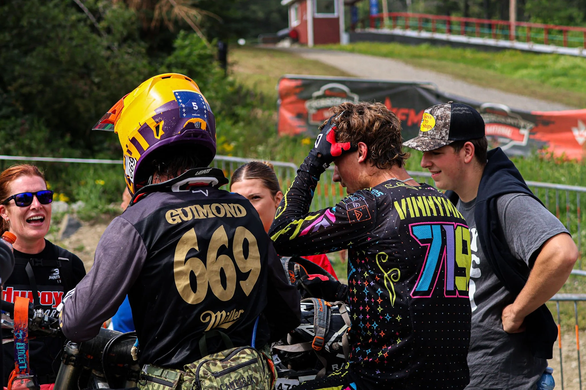 Group of motocross racers and supporters outdoors, one racer with a yellow, purple, and orange helmet, surrounded by smiling people, with a red building and trees in the background.