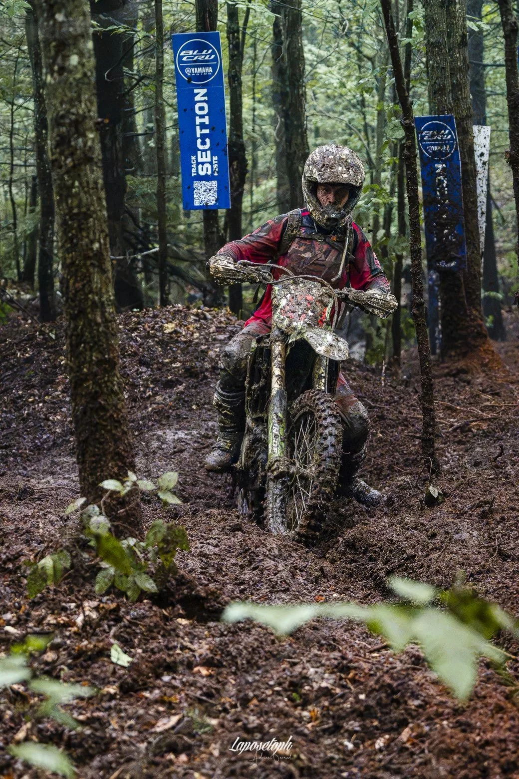 A person riding a dirt bike through a muddy forest trail, wearing a helmet and red gear, with blue 'Yamaha' signs in the background.