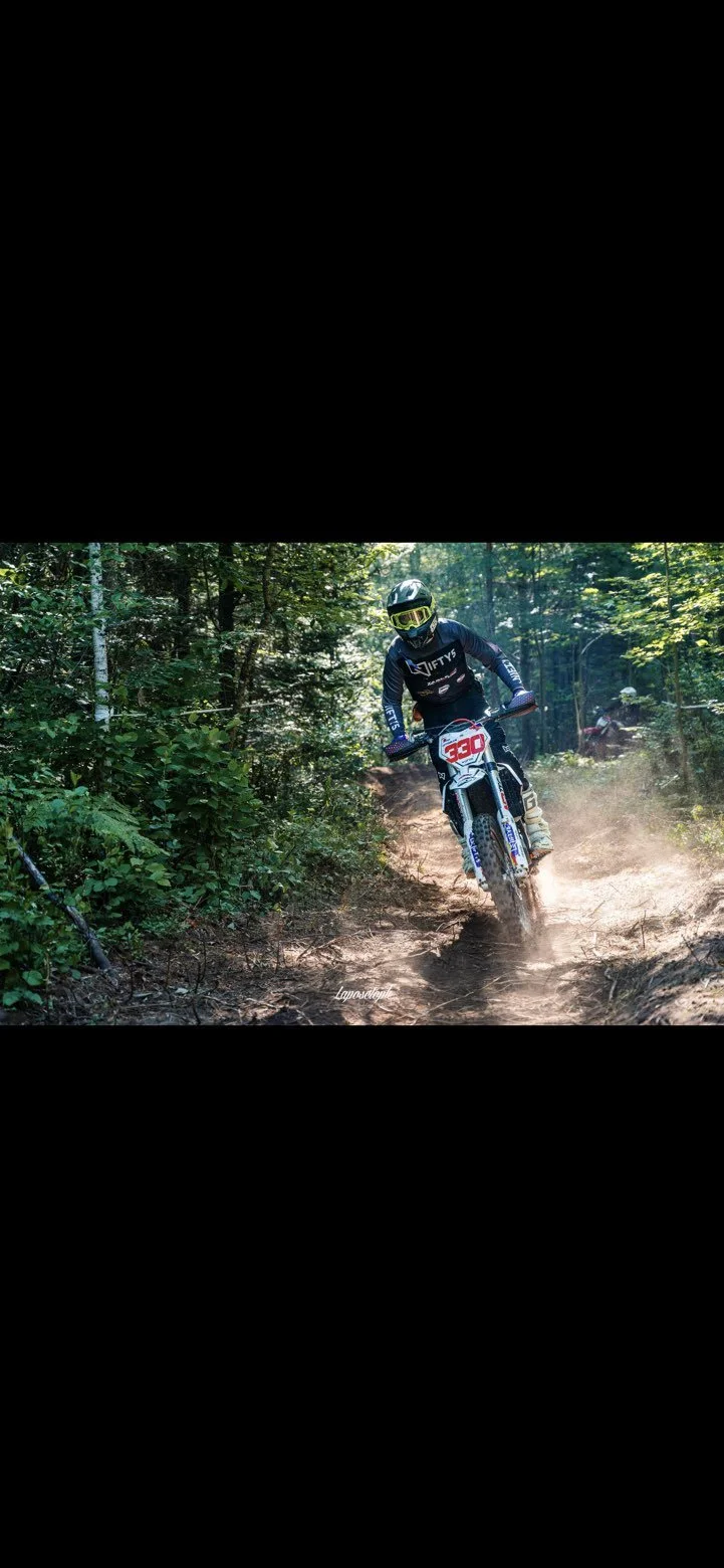 Motocross rider wearing helmet and gear riding through a forest trail, kicking up dust.