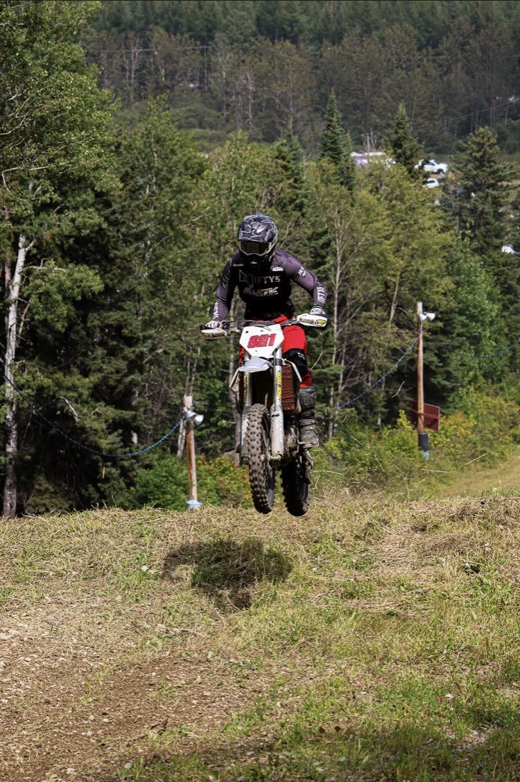 A person in a black helmet and riding gear is jumping on a dirt bike on a trail surrounded by trees.