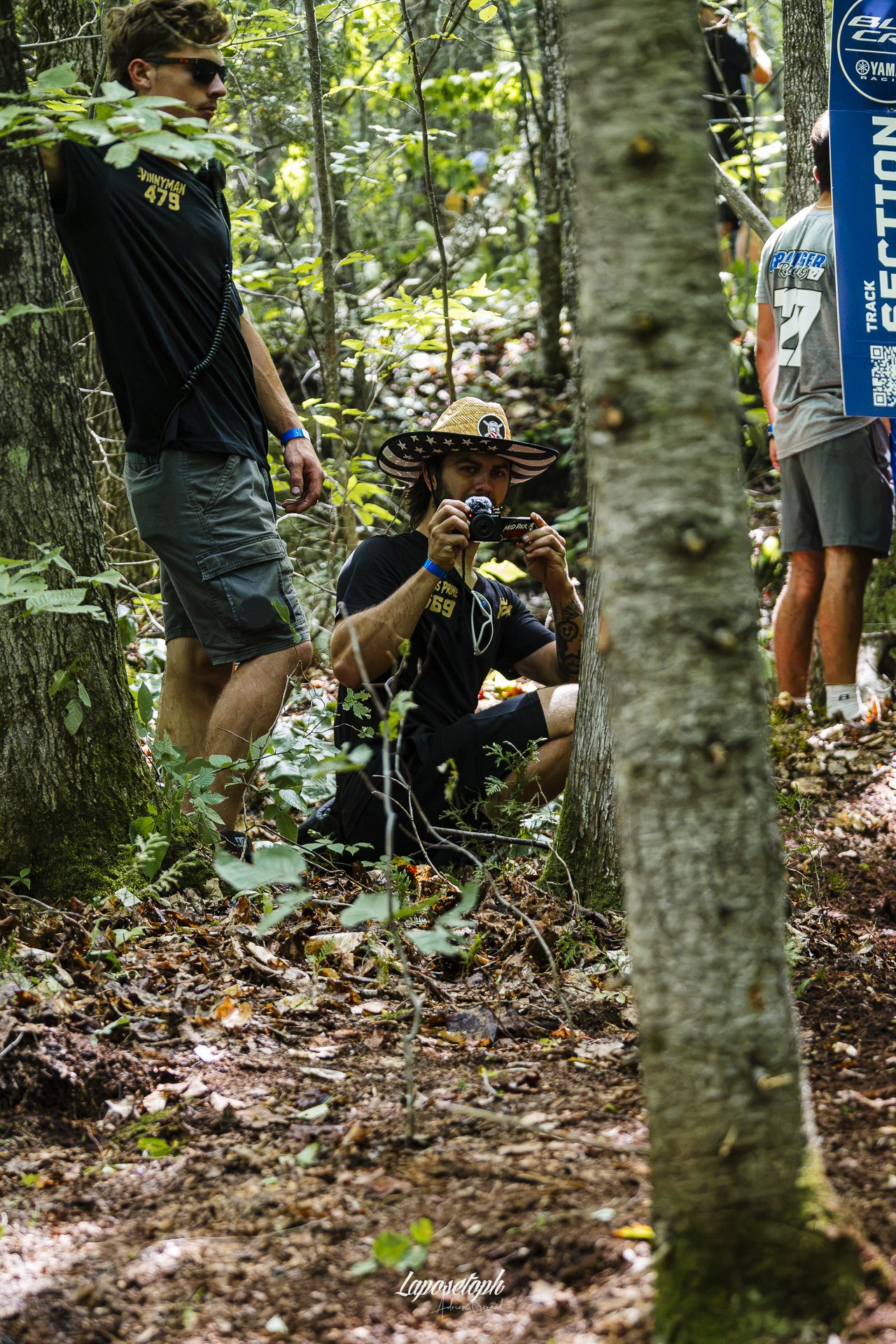 A person wearing a sombrero hat and black T-shirt sitting in a wooded area taking a photo with a camera, surrounded by trees and leaves on the ground, with two other people standing nearby.