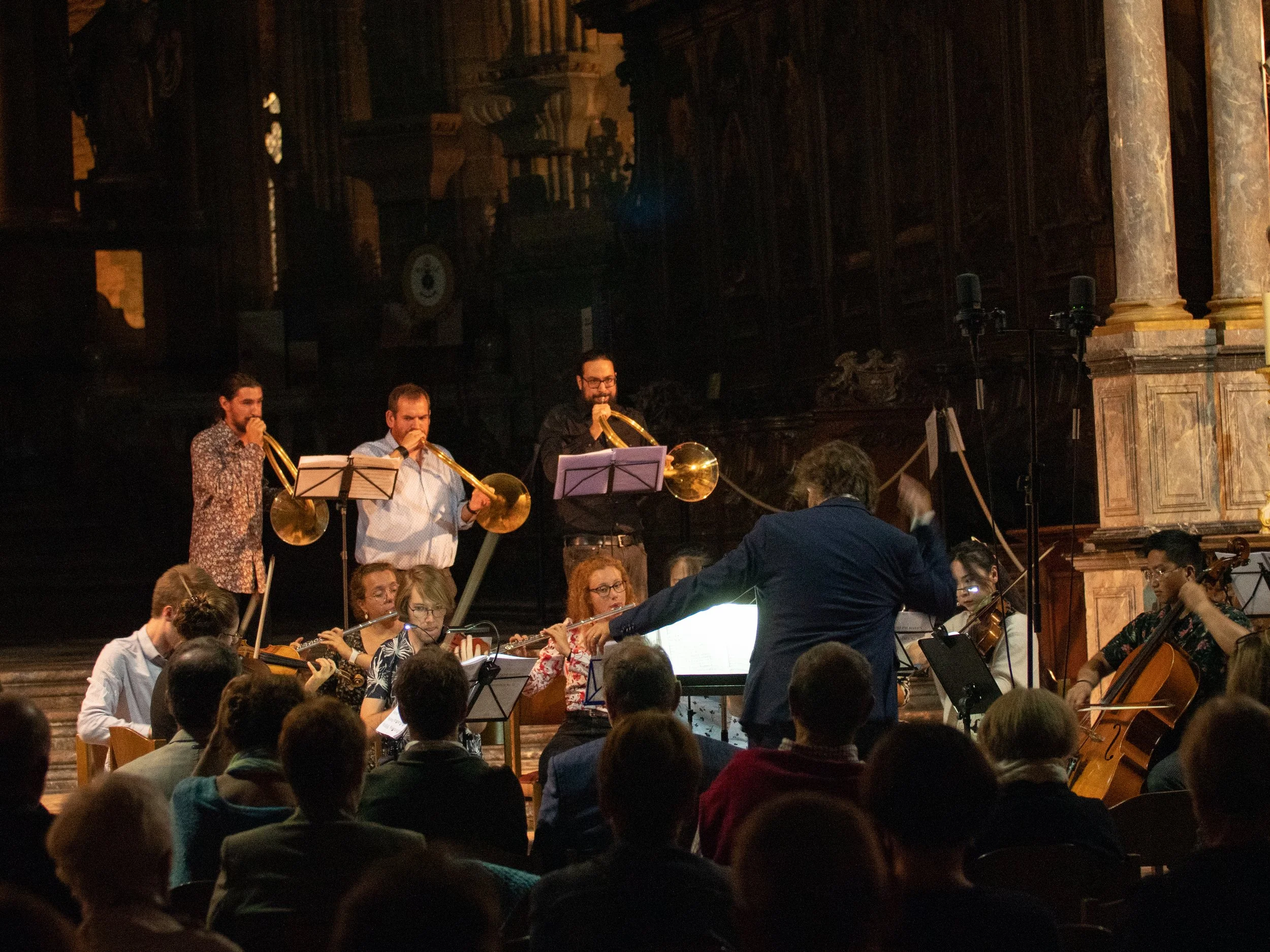 Un orchestre joue dans une salle ornée de décorations anciennes, avec des musiciens et un chef d'orchestre en avant, entourés d'un public.
