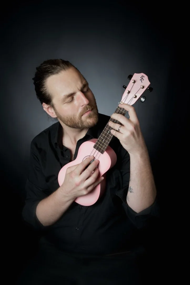 Un homme avec les yeux fermés joue de la guitare ukulélé rose contre un fond sombre.