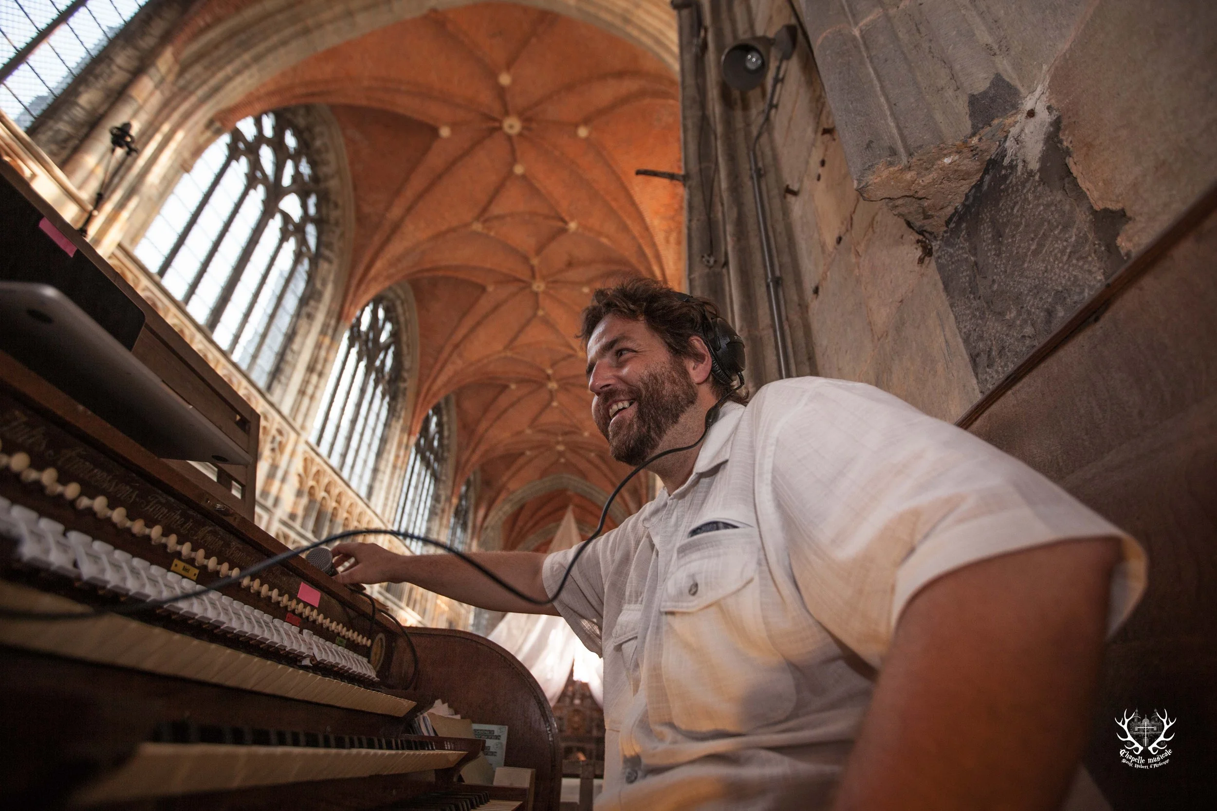 Un homme souriant joue du piano dans une cathédrale avec une voûte en brique et de grands vitraux derrière lui.