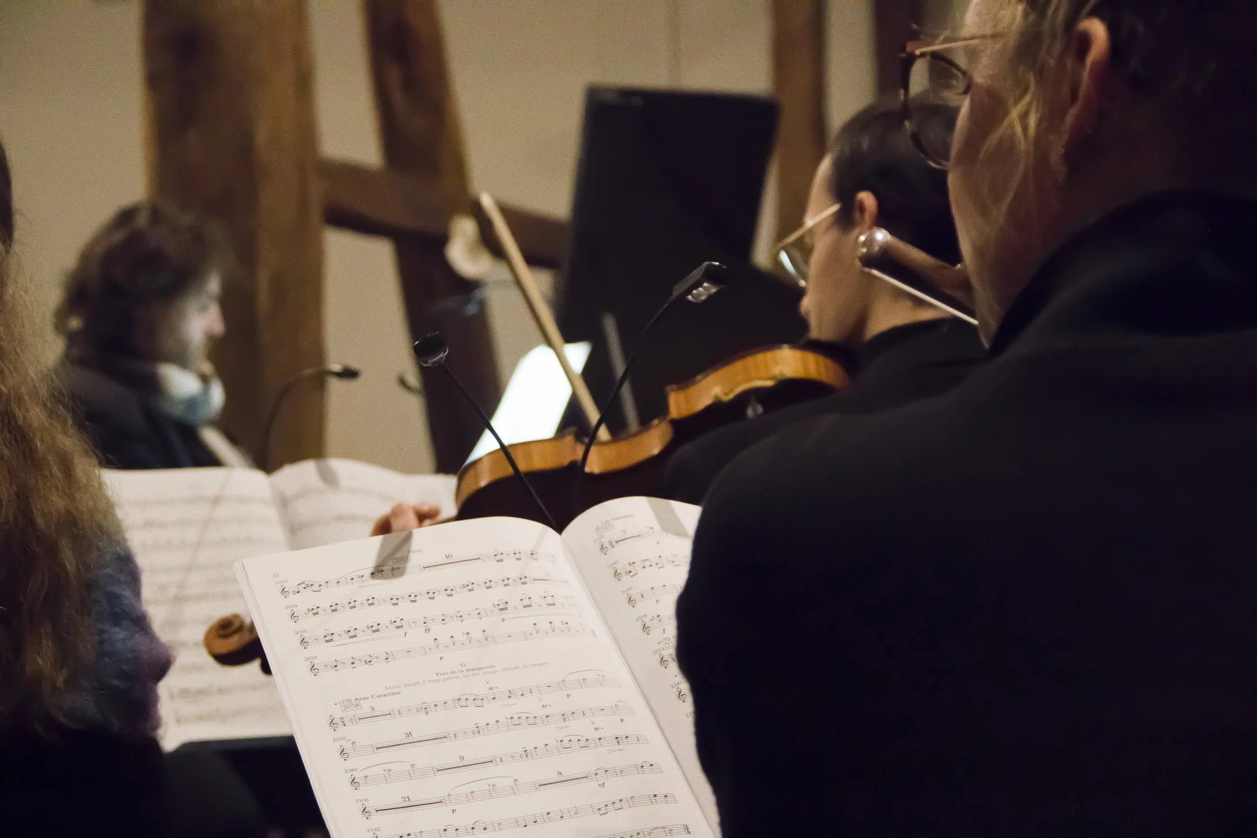 Groupe de musiciens jouant du violon lors d'une répétition ou d'un concert, avec des partitions sur leur partition. L'image montre plusieurs personnes concentrées, certaines portant des lunettes, dans un environnement intérieur.