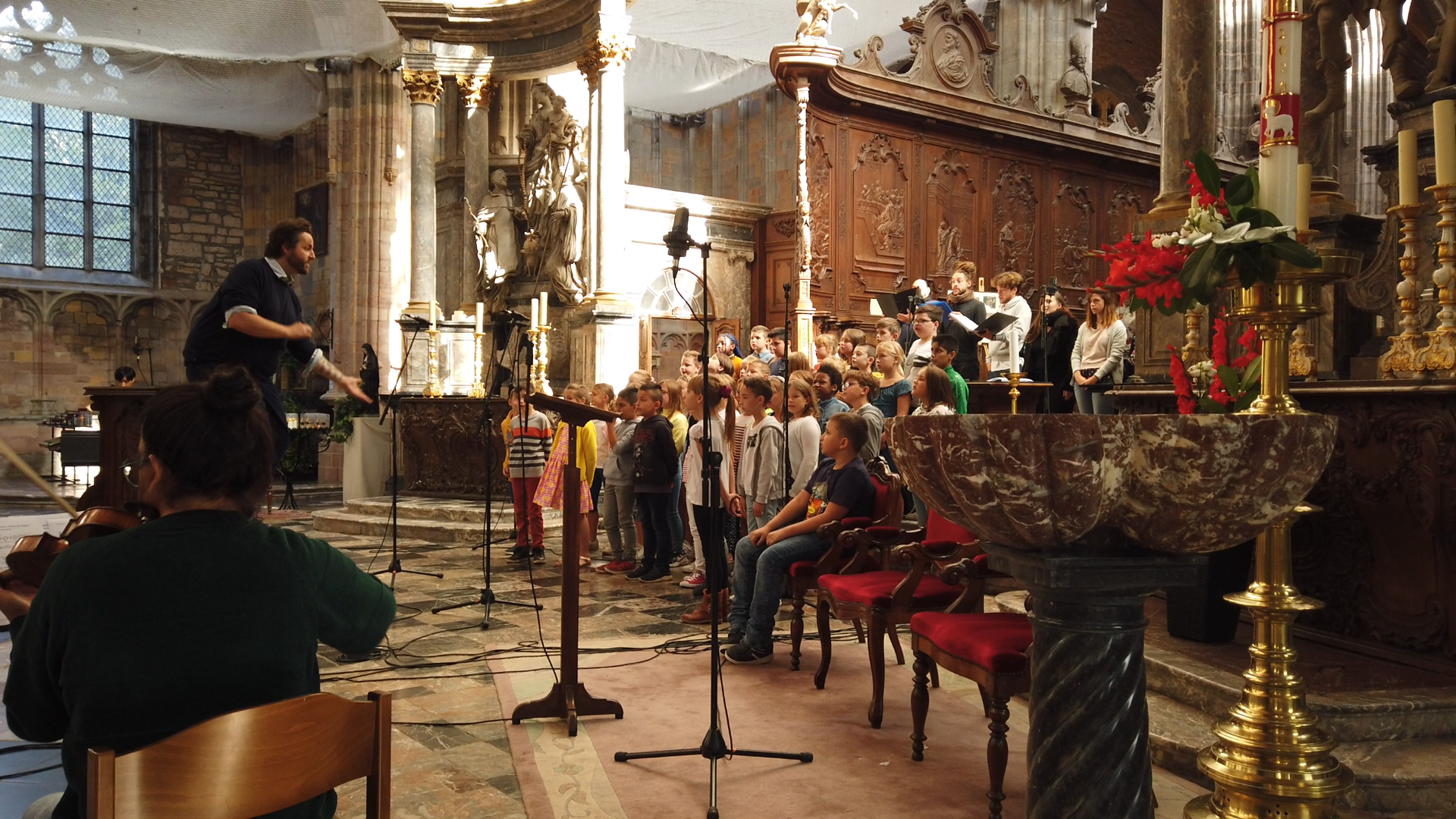 Groupe d'enfants chantant dans une église, dirigés par un homme, avec un orchestre et un chœur d'adultes en arrière-plan, décorée avec des statues, des candélabres dorés et des vases de fleurs roses et blanches.