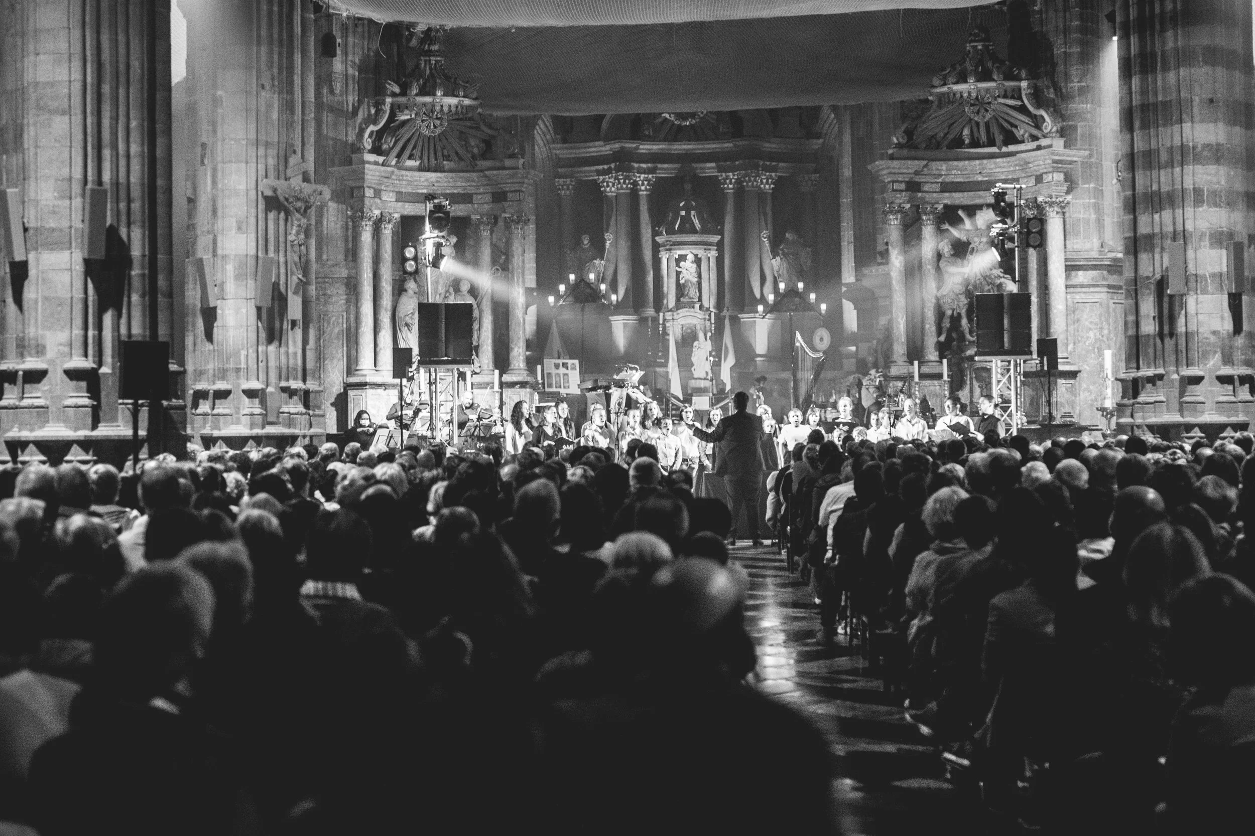 Un chœur de personnes chantant dans une église gothique avec des colonnes ornées et des statues, éclairé par des projecteurs, lors d'une performance musicale.