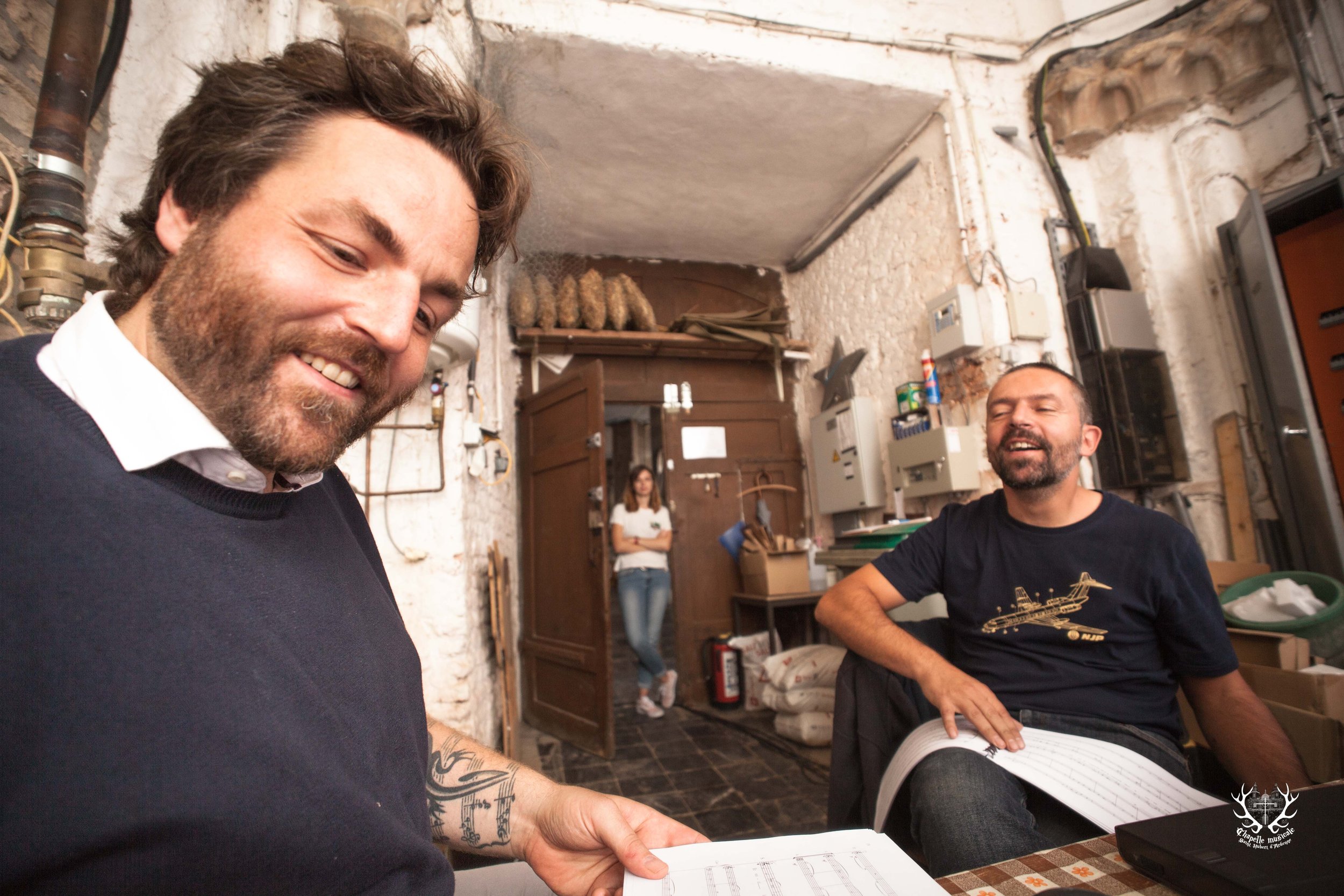 Deux hommes souriants dans une pièce aux murs en briques, l'un tenant une feuille de papier avec des notes, l'autre assis à une table. Une femme en arrière-plan.