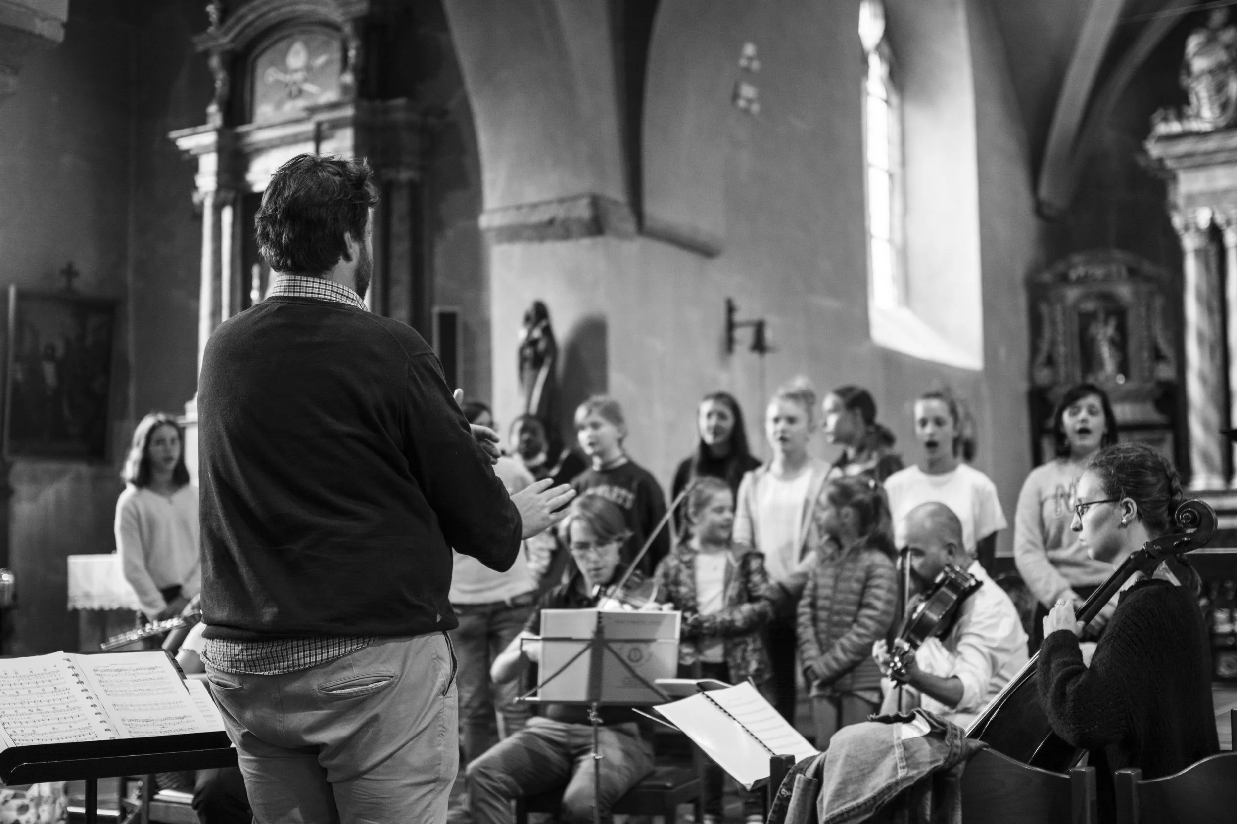 Un homme qui dirige une pratique vocale ou chorale dans une église, entouré de plusieurs jeunes chanteurs et musiciens, dans une église avec des vitraux et des décorations religieuses.