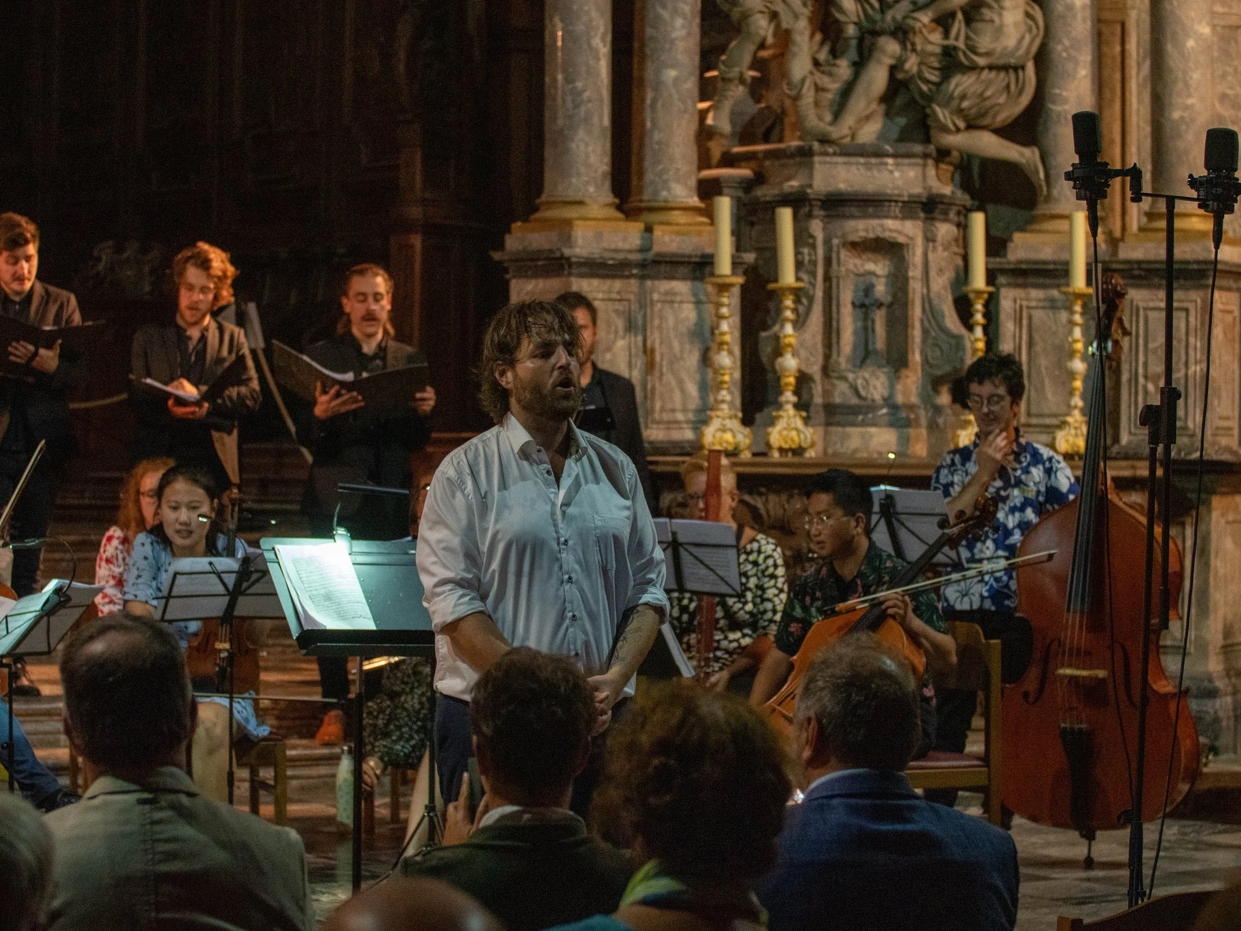 Une performance musicale dans une église avec un chœur de chanteurs, un chef d'orchestre masculin au centre, entouré d'instrumentistes dont une violoniste. L'église est ornée de colonnes et d'une grand sculpture en arrière-plan.