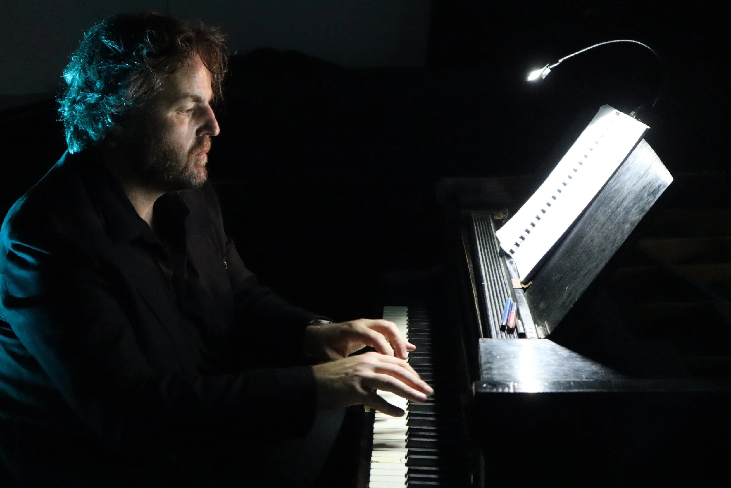 Homme jouant du piano dans une pièce sombre, éclairé par la lumière de la partition.