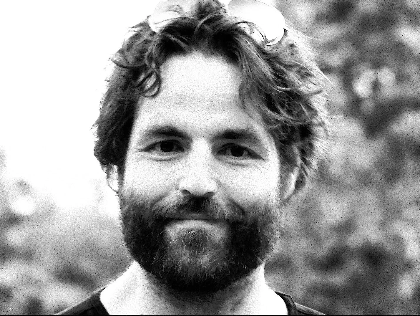 Portrait d'un homme avec une barbe et des cheveux bouclés, souriant légèrement, en extérieur.