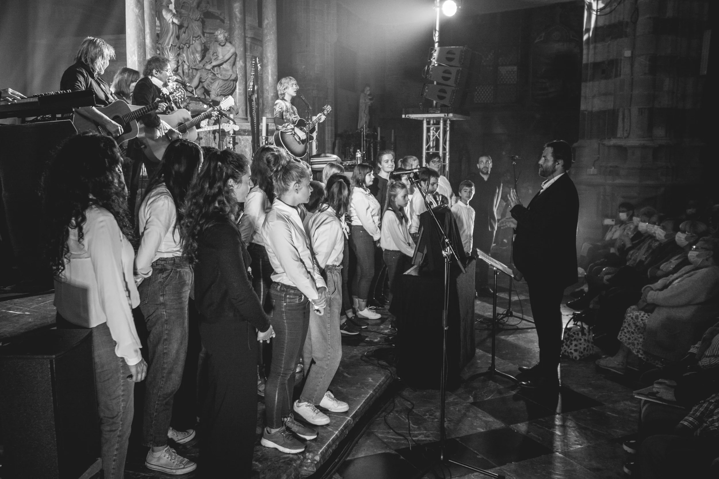 Un groupe de chanteurs, principalement des jeunes, se tient en formations lors d'une performance ou d'une répétition, sous la direction d'un chef de chœur, dans un environnement intérieur avec un décor artistique en arrière-plan.