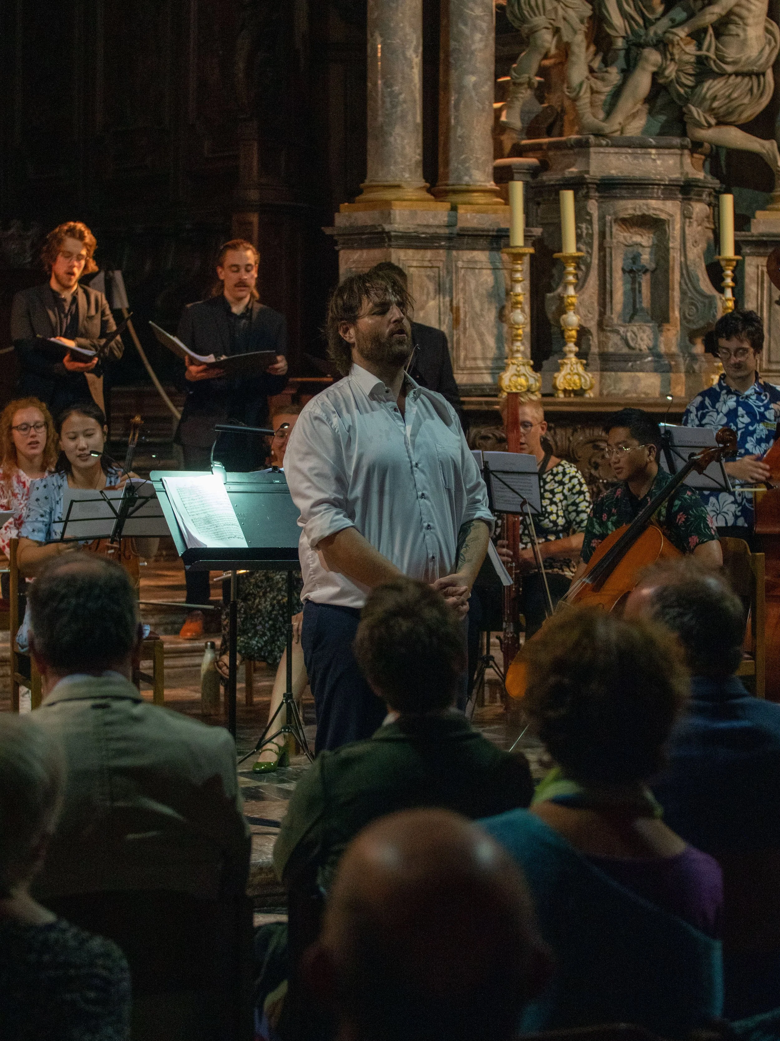 Un groupe de musiciens et un chef d'orchestre en train de jouer dans une église ou un lieu historique avec des colonnes en marbre, des bougies et une statue sur l'autel.