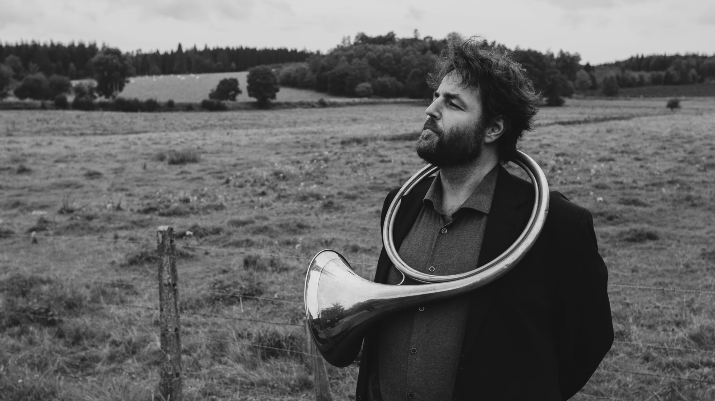 Homme avec barbe et cheveux en désordre portant un saxophone autour du cou, regardant vers l'horizon dans un paysage de campagne avec arbres et champs en arrière-plan, image en noir et blanc.
