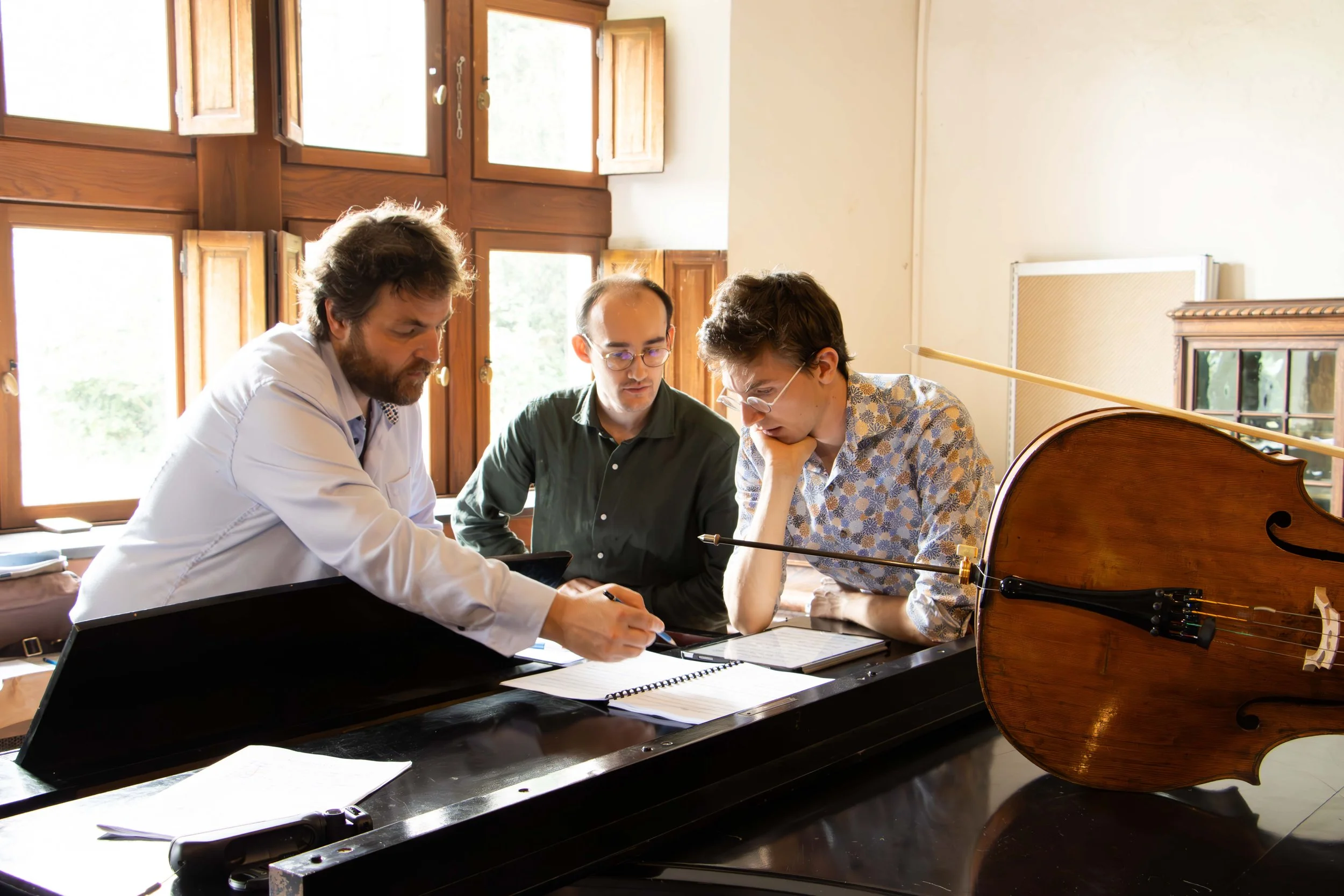Trois hommes autour d'un piano, concentrés sur une partition, avec un violon posé en évidence. La scène se déroule dans une pièce avec de grandes fenêtres en bois.