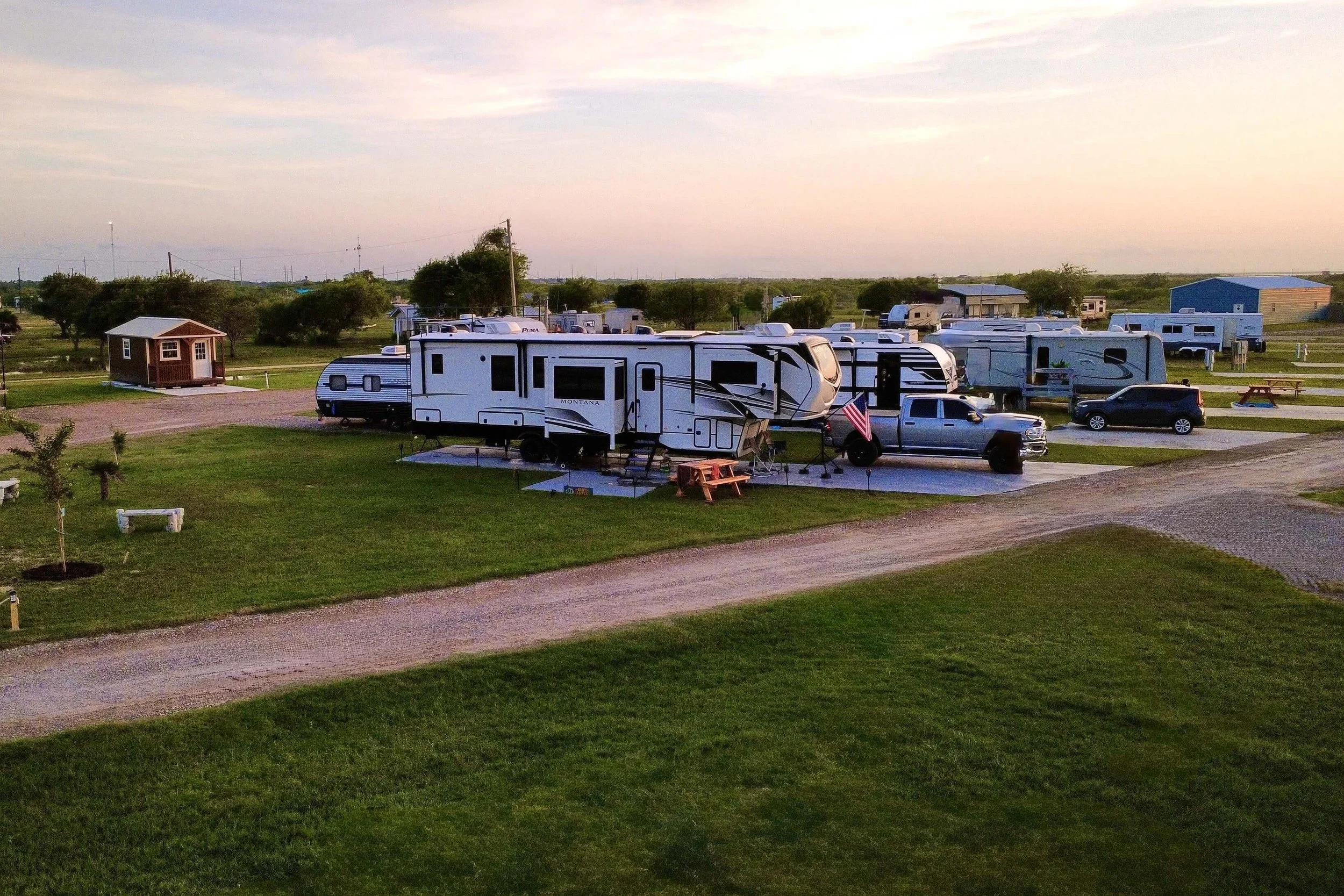 A campground with multiple RVs, a small brown shed, and picnic tables on a grassy field, sunset in the background.