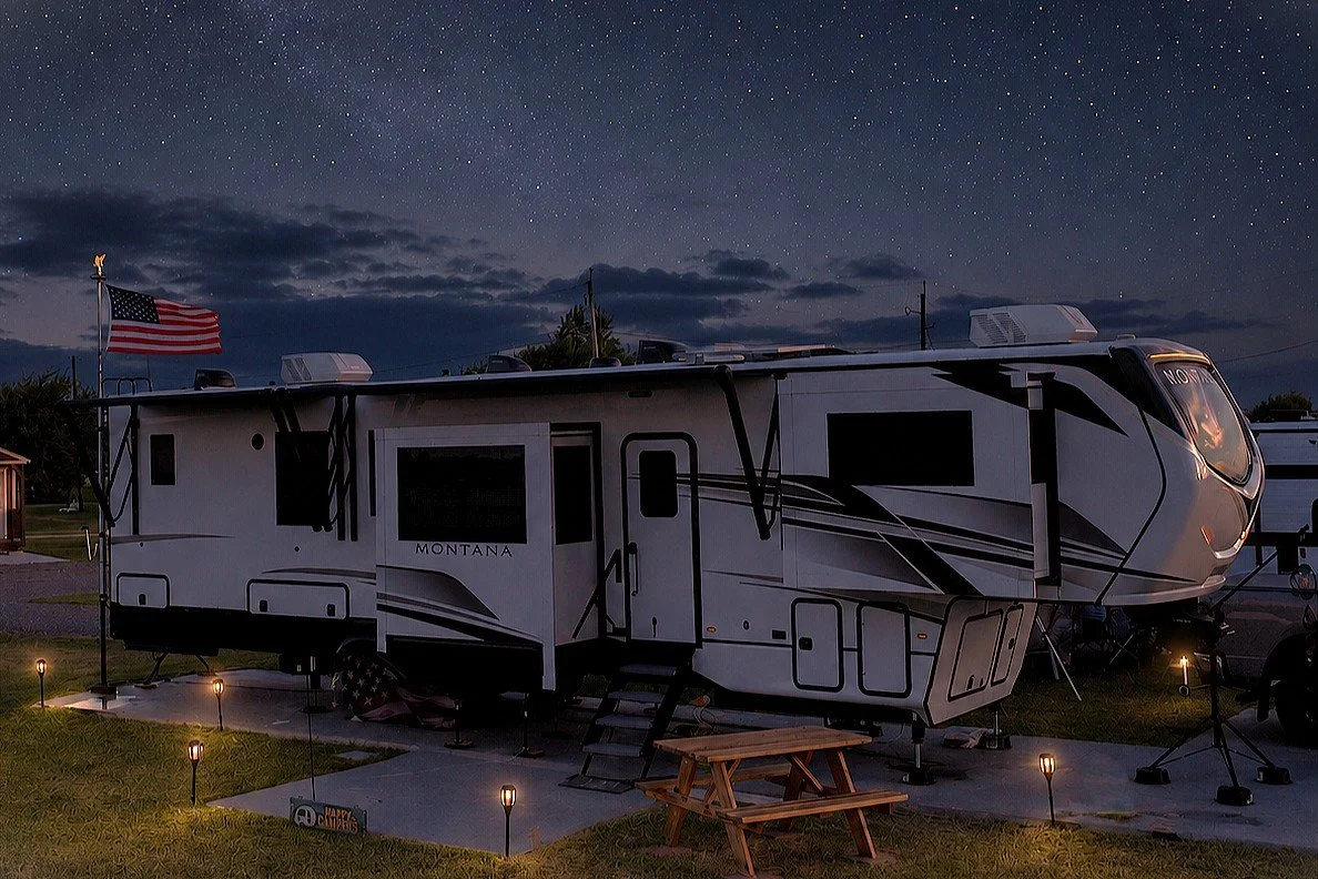 A large white and black Montana RV truck with a slide-out section, parked outdoors at night under a starry sky. An American flag is flying on a pole nearby, and small lights illuminate the area. A picnic table is in foreground.
