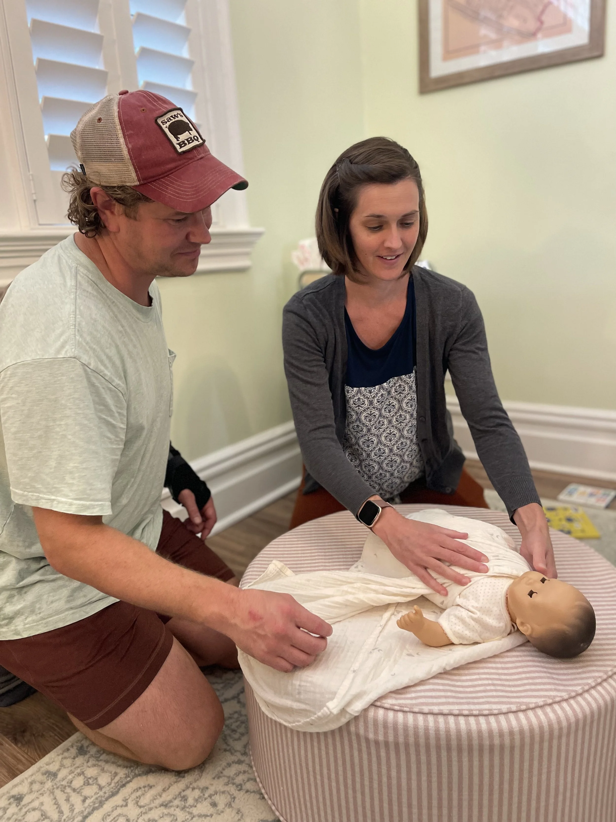 Elissa is knelt showing a man how to swaddle a baby using a doll.