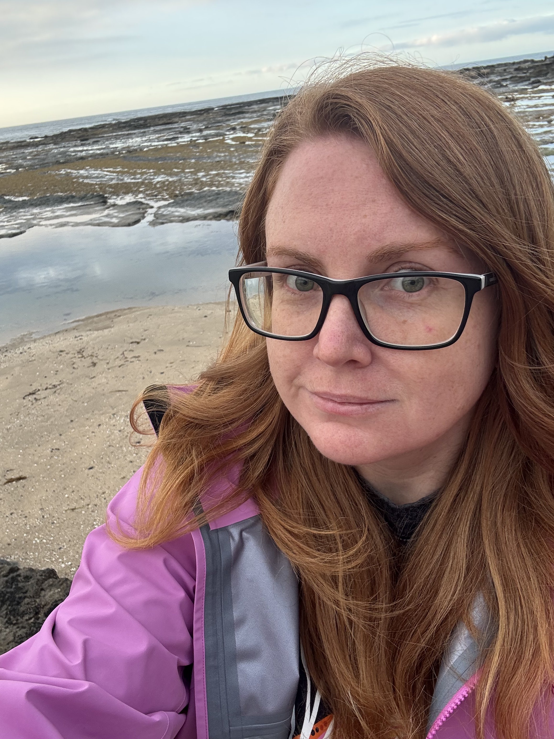 Photo of Meg who has long red hair and glasses wearing a pink raincoat at a beach in Naarm.