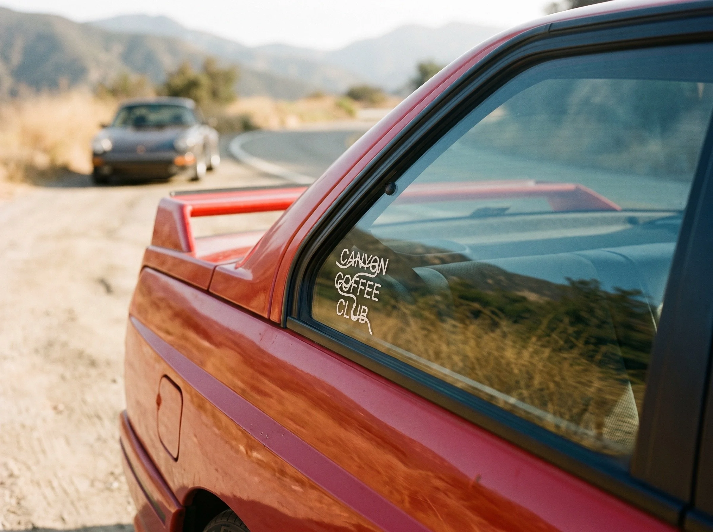 Close-up of a red vehicle with a sticker on the window that reads 'Canyon Coffee Club'. In the background, there is a blurred view of another vehicle and mountainous terrain.