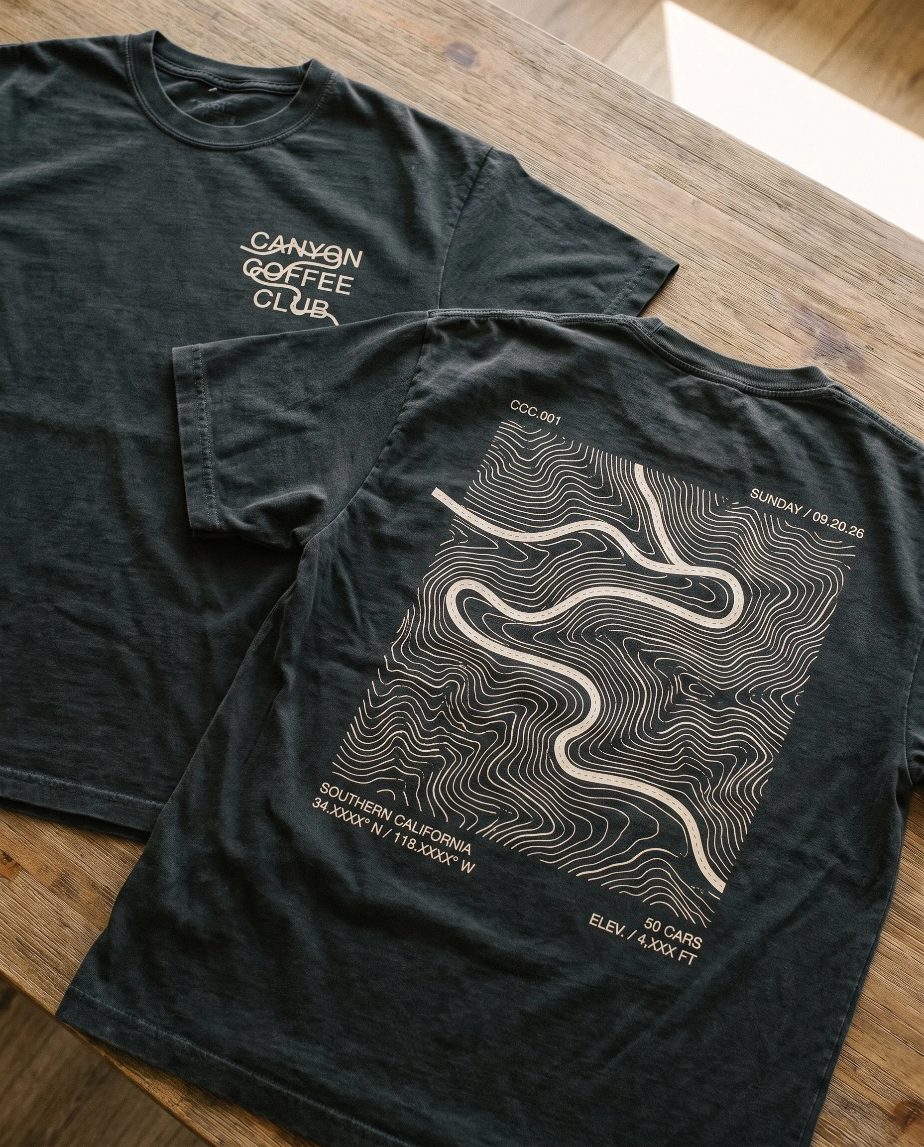 Black T-shirt with map design and white text, lying on a wooden table. The shirt features topography lines and coordinates, referencing Southern California, with details like elevation and car count.