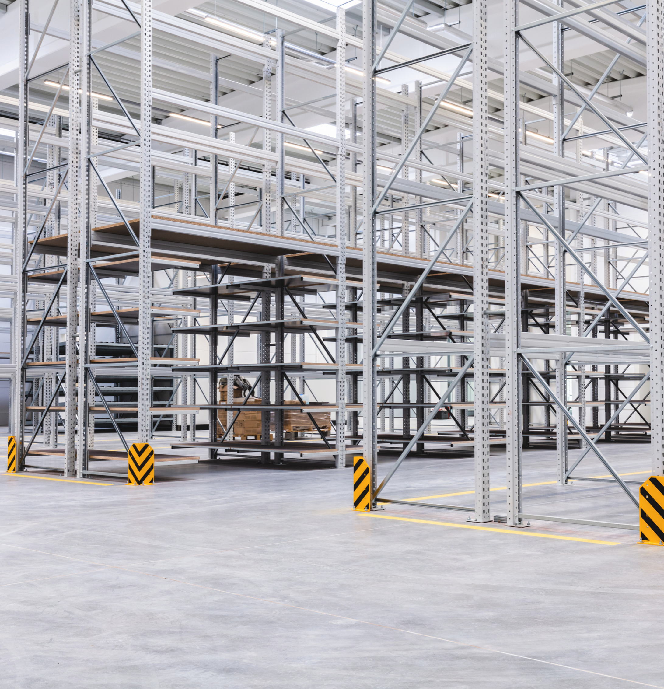 Empty warehouse with tall metal shelving units, some with wooden pallets and boxes, yellow and black safety barriers, and a concrete floor with marked lines.