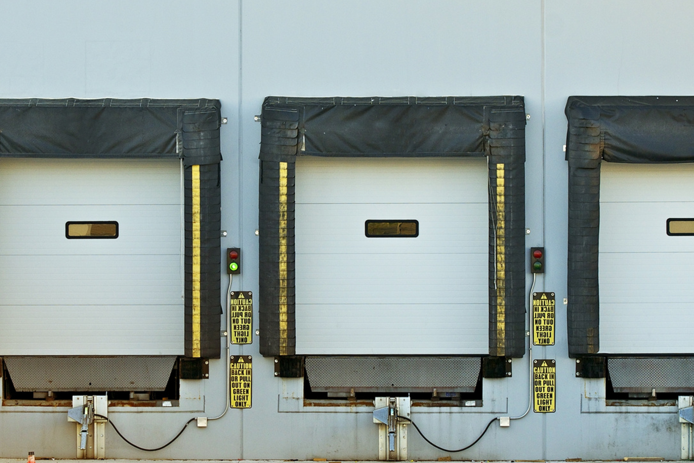 Three loading dock doors with black retractable bumpers and yellow caution signs, one door with a green light, in an industrial setting.
