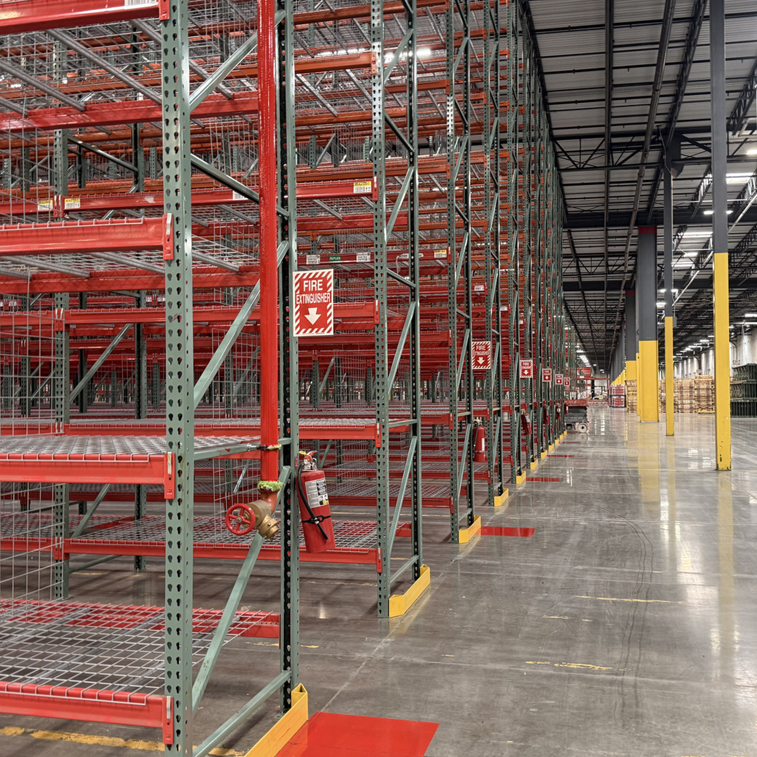 Empty industrial warehouse with tall metal shelving units, fire extinguisher, and fire alarm, with a polished concrete floor and overhead lighting.