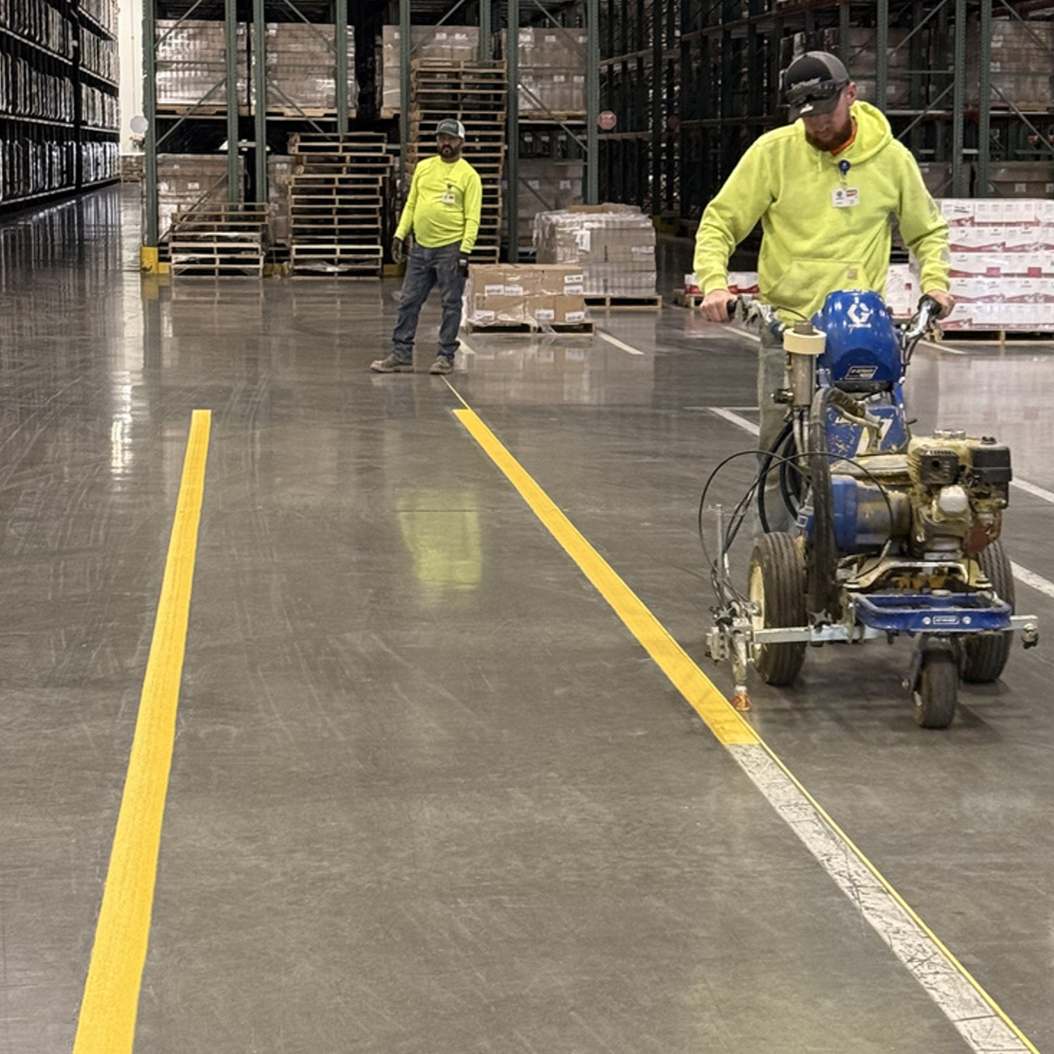 Two workers in high-visibility yellow jackets working in a warehouse with shelves and pallets in the background. One worker is standing, and the other is operating a floor marking machine that applies yellow lines on the polished concrete floor.