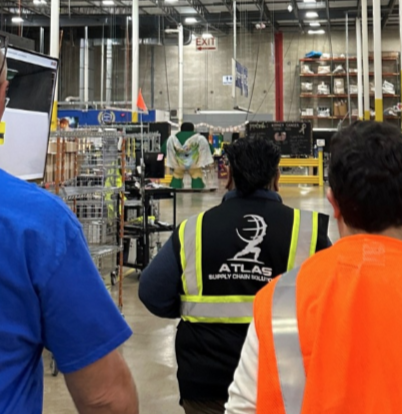 Employees in safety vests and uniform standing in an industrial warehouse or supply center with shelves and equipment.