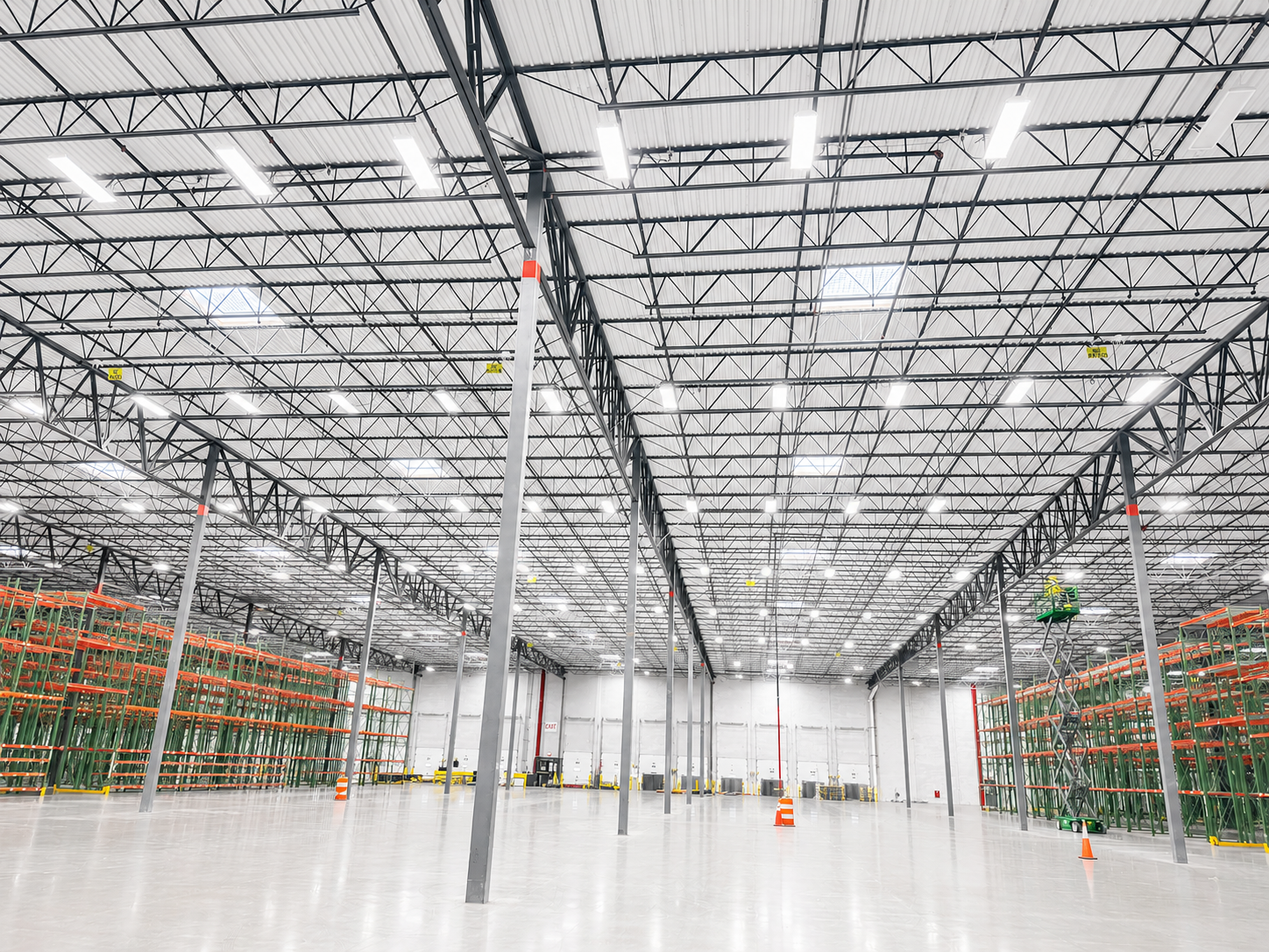 A spacious, empty warehouse with a high ceiling, metal beams, bright overhead lighting, and storage racks on the sides.
