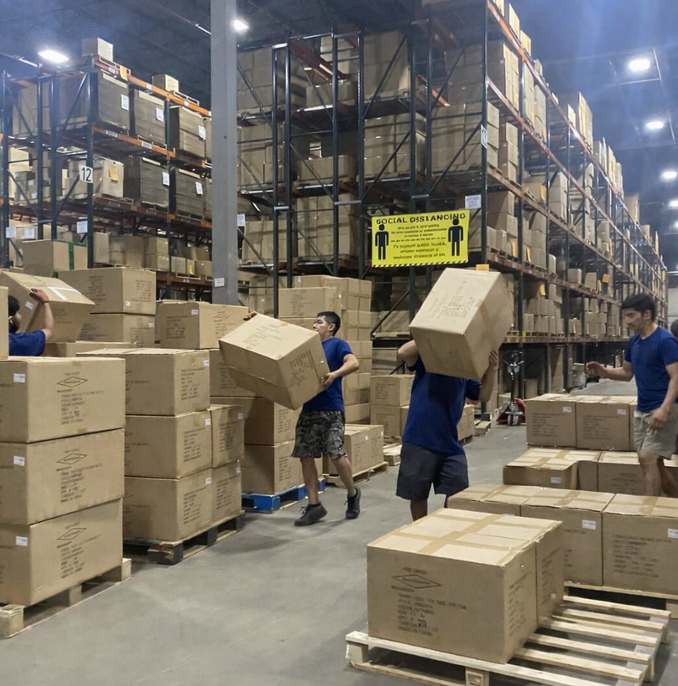 Workers at a warehouse unloading boxed goods from pallets in an industrial setting with tall shelving and bright overhead lighting.
