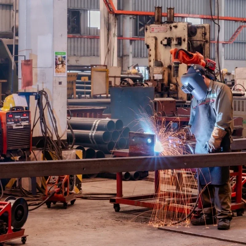A worker wearing a welding helmet and protective gloves welding metal in an industrial workshop, with sparks flying around.