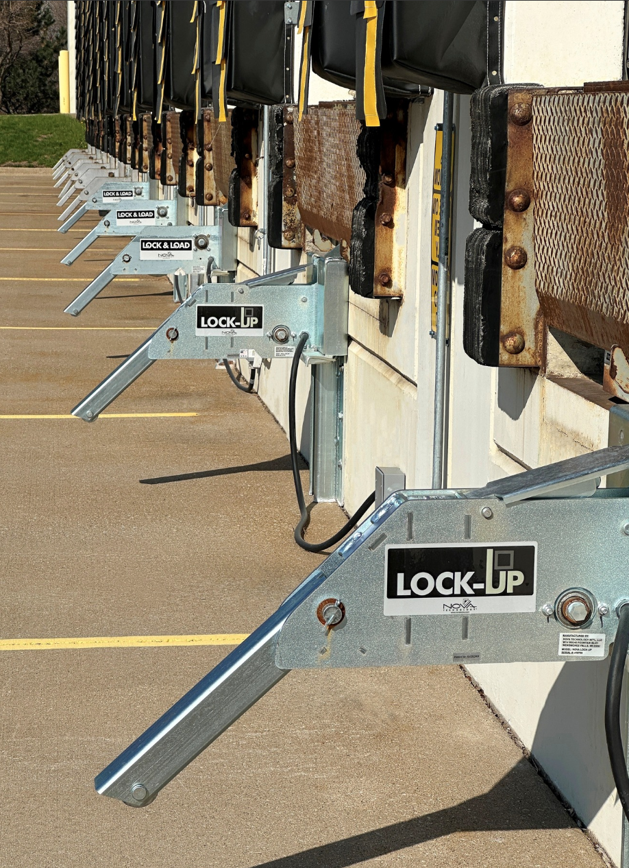 Several industrial jersey barriers with metal locking mechanisms, lined up along a concrete sidewalk, with arrow handles on the locks and yellow straps attached to black bags on top.